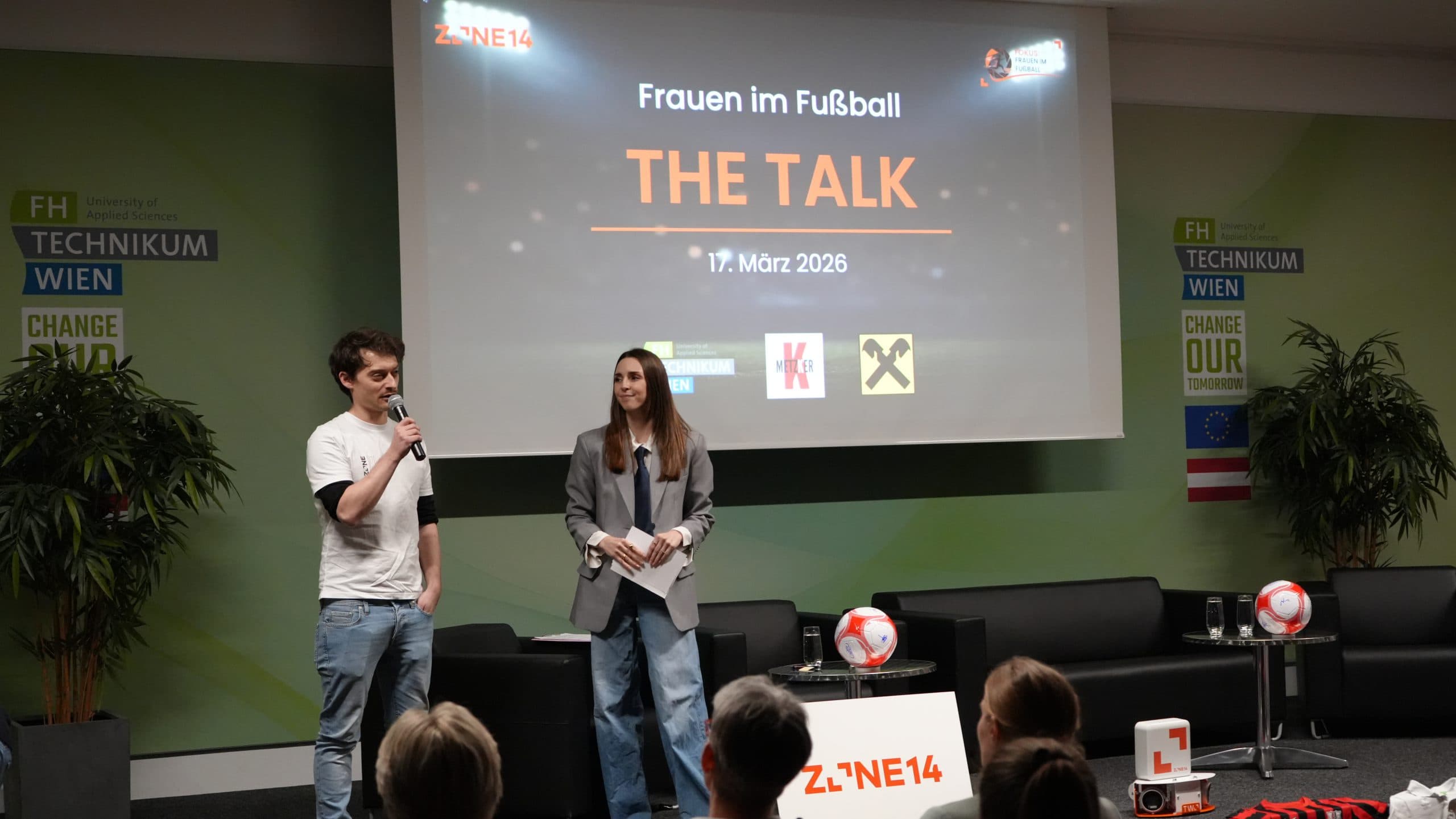 Zwei Moderatorinnen stehen mit Mikrofonen auf der Bühne vor einem Publikum. Hinter ihnen steht auf einer Leinwand Frauen im Fußball THE TALK 17. März 2026. Fußbälle und Transparente schmücken die Bühne.