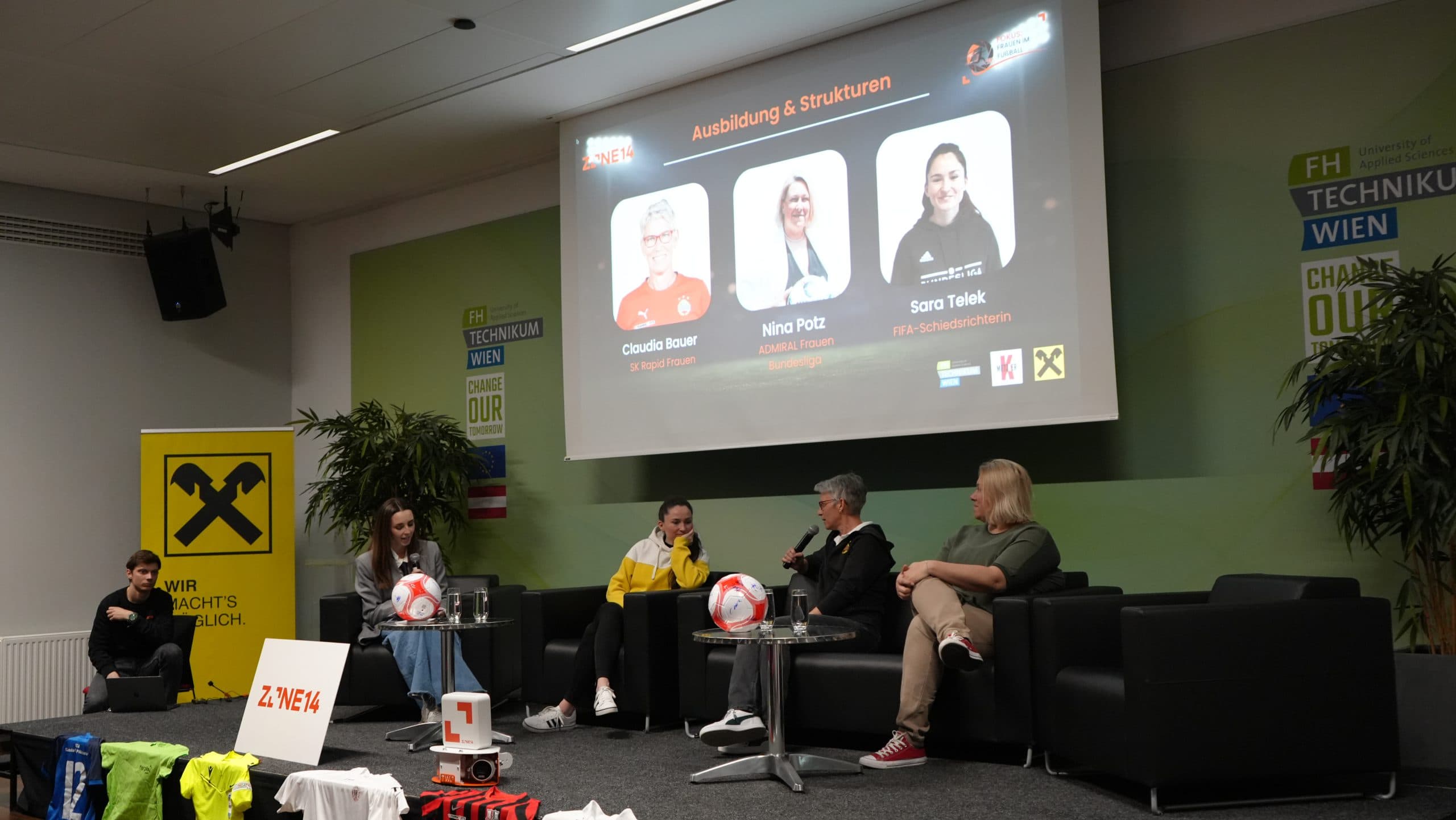 Vier Frauen sitzen auf einer Bühne und führen eine Podiumsdiskussion. Hinter ihnen ist ein Bildschirm mit ihren Fotos und Namen sowie Logos zu sehen. Vor der Bühne sind Sporttrikots und ein Fußball ausgestellt.