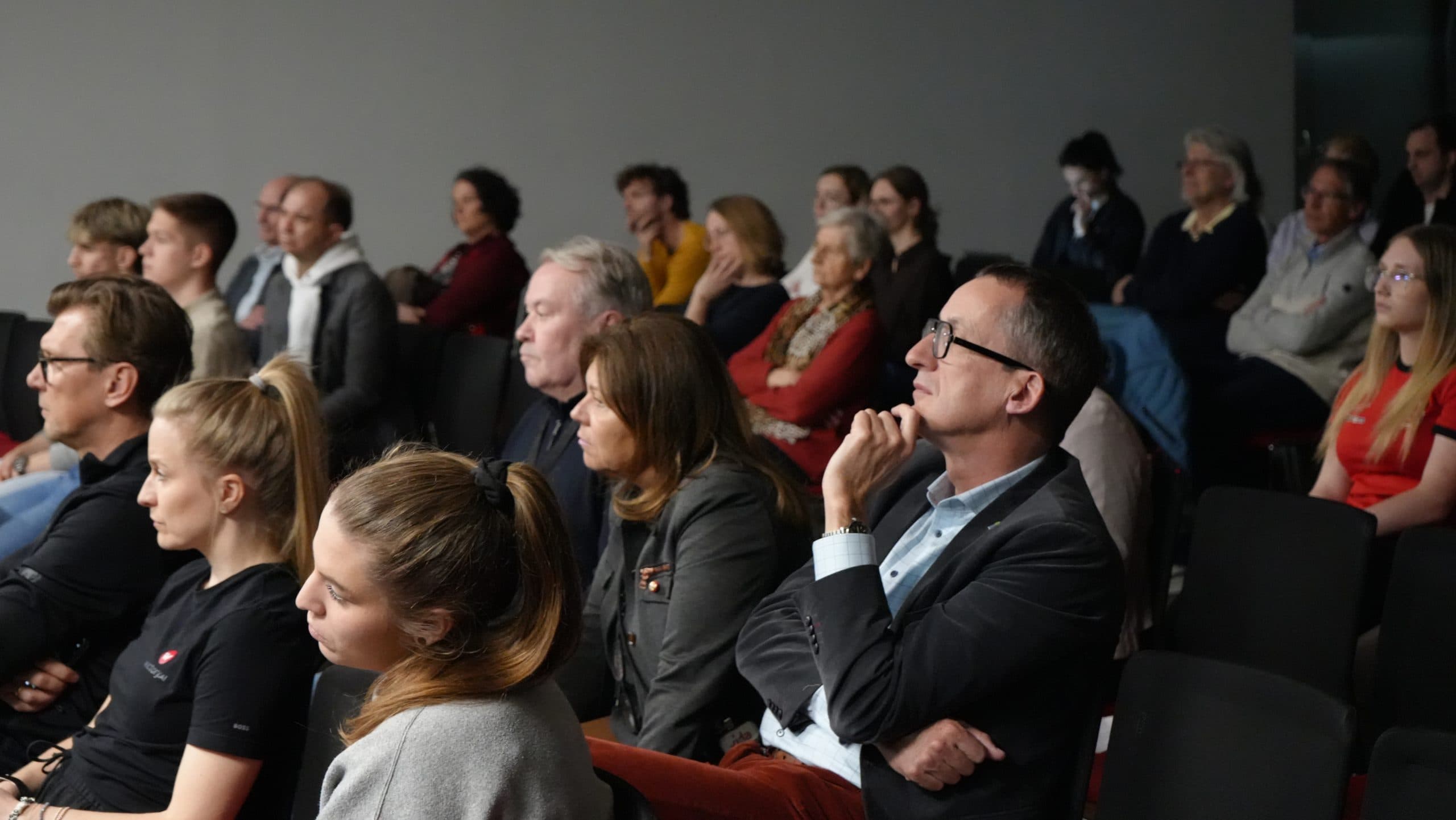 Eine Gruppe von Menschen unterschiedlichen Alters sitzt aufmerksam in Reihen schwarzer Stühle und beobachtet in einem gut beleuchteten Raum etwas Unsichtbares. Die meisten scheinen konzentriert und beschäftigt zu sein, einige stützen ihr Kinn auf ihre Hände.