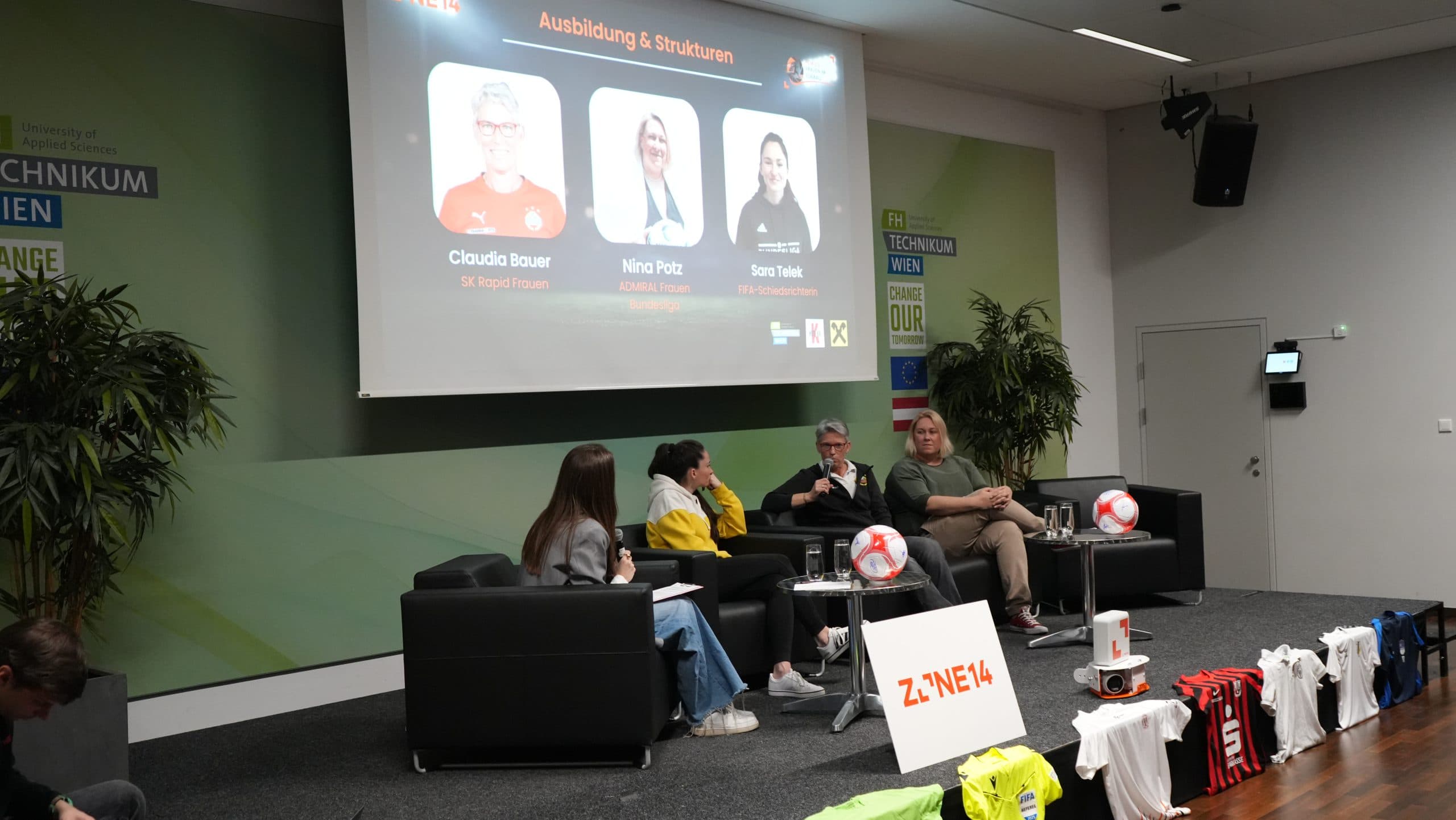 Drei Diskussionsteilnehmer und ein Moderator sitzen auf der Bühne in Sesseln, hinter denen ein Bildschirm ihre Namen und Fotos zeigt. Vorne sind Fußballtrikots und -bälle ausgestellt, und die Umgebung scheint eine Konferenz oder ein Seminar zu sein.