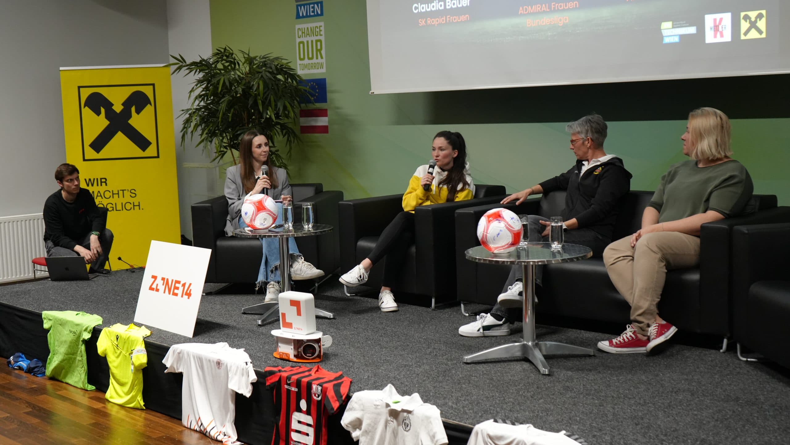 Vier Personen sitzen bei einer Podiumsdiskussion auf einer Bühne, vor ihnen liegen Fußbälle und Trikots. Ein Moderator hält ein Mikrofon, während die anderen zuhören. Im Hintergrund sind ein 'Wir machts möglich'-Banner und ein digitaler Bildschirm zu sehen.