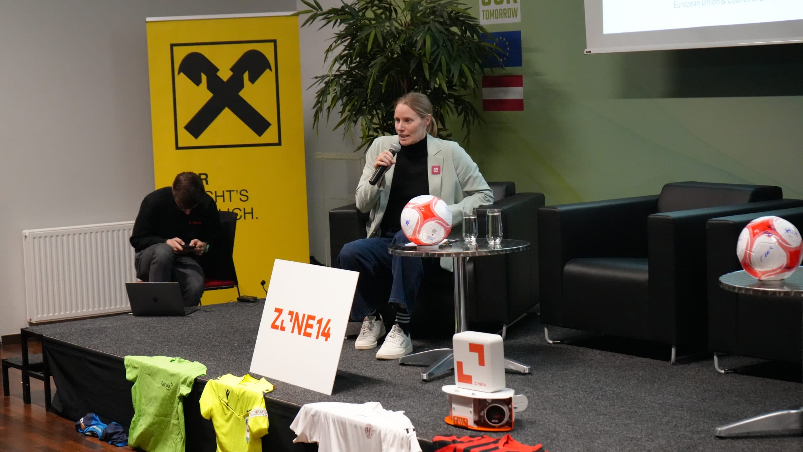 Eine Frau sitzt auf einer Bühne und hält ein Mikrofon und einen Fußball in der Hand. Um sie herum sind Sporttrikots, Fußbälle und Ausrüstungsgegenstände ausgestellt. Ein Mann sitzt in der Nähe auf dem Boden und benutzt einen Laptop. Im Hintergrund ist ein großes gelbes Transparent zu sehen.