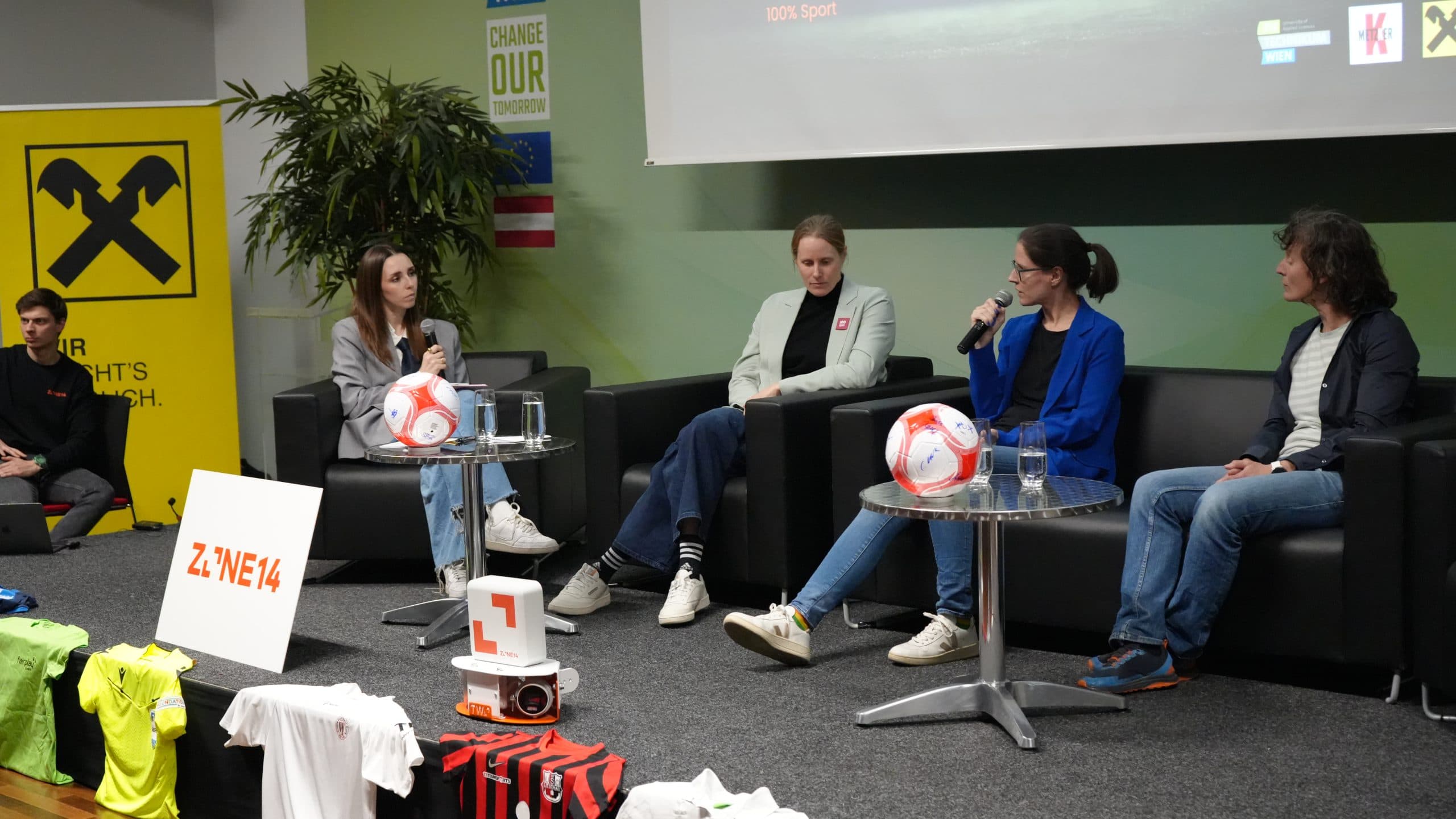 Vier Frauen sitzen auf einem Podium und diskutieren mit Mikrofonen in der Hand. Um sie herum sind Fußbälle, Sporttrikots und Markenbanner zu sehen. Im Hintergrund sind ein Bildschirm und eine Pflanze zu sehen.