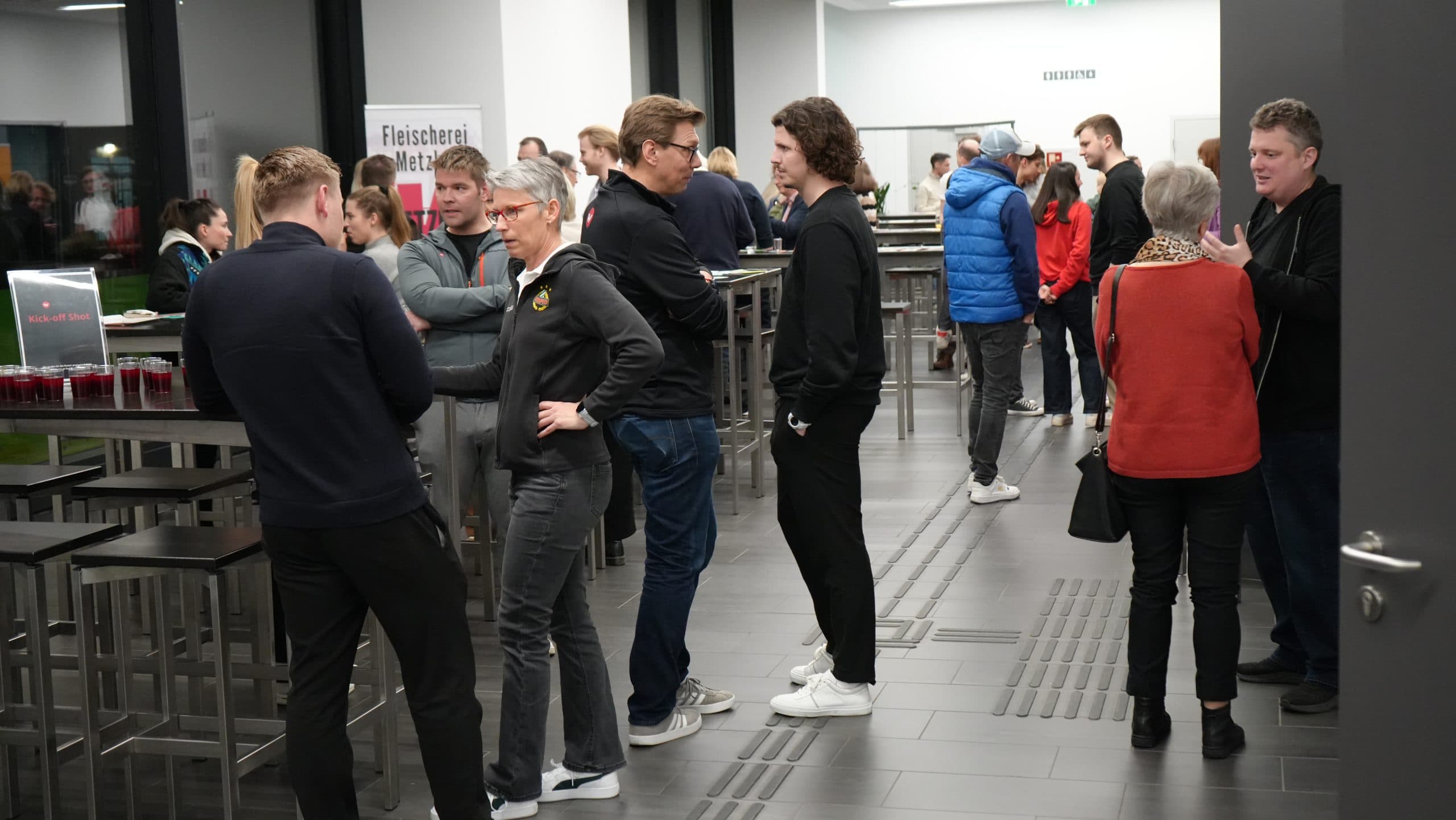 Eine Gruppe von Menschen steht und unterhält sich in einem modernen Innenraum mit Stehtischen und Hockern. Einige halten Getränke in der Hand, und auf einem Tisch im Hintergrund stehen Schilder und Getränke. Die Atmosphäre wirkt locker und gesellig.