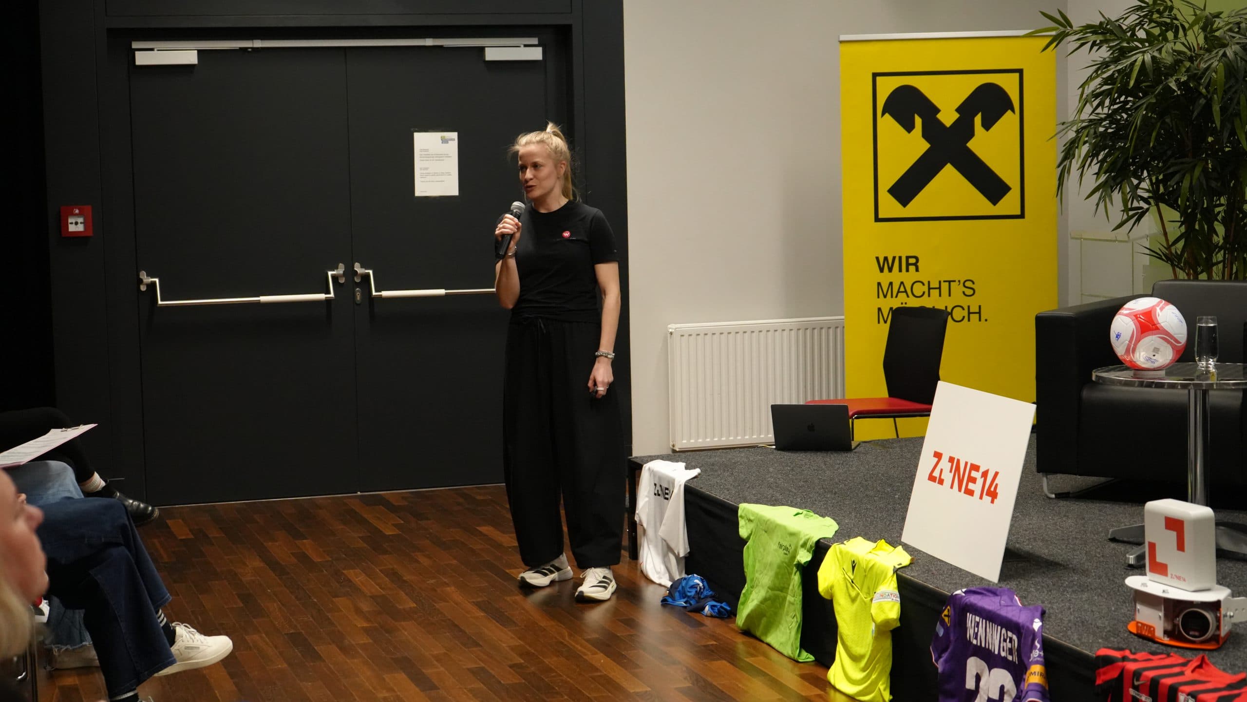 Eine Frau spricht in ein Mikrofon auf der Bühne neben einer Auslage mit Sporttrikots, einem Fußball und einer gelben Raiffeisen-Fahne. Im Publikum sitzen mehrere Personen, und in der Nähe stehen eine Pflanze und ein schwarzer Sessel.