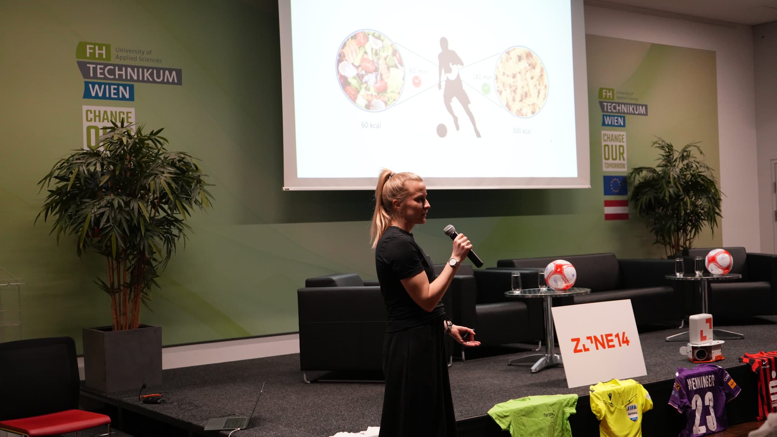 Eine Frau mit einem Mikrofon präsentiert auf der Bühne vor einem Bildschirm, der eine Folie zum Thema Ernährung zeigt. Sporttrikots und Fußbälle sind zu sehen. Im Hintergrund steht 'FH Technikum Wien'.