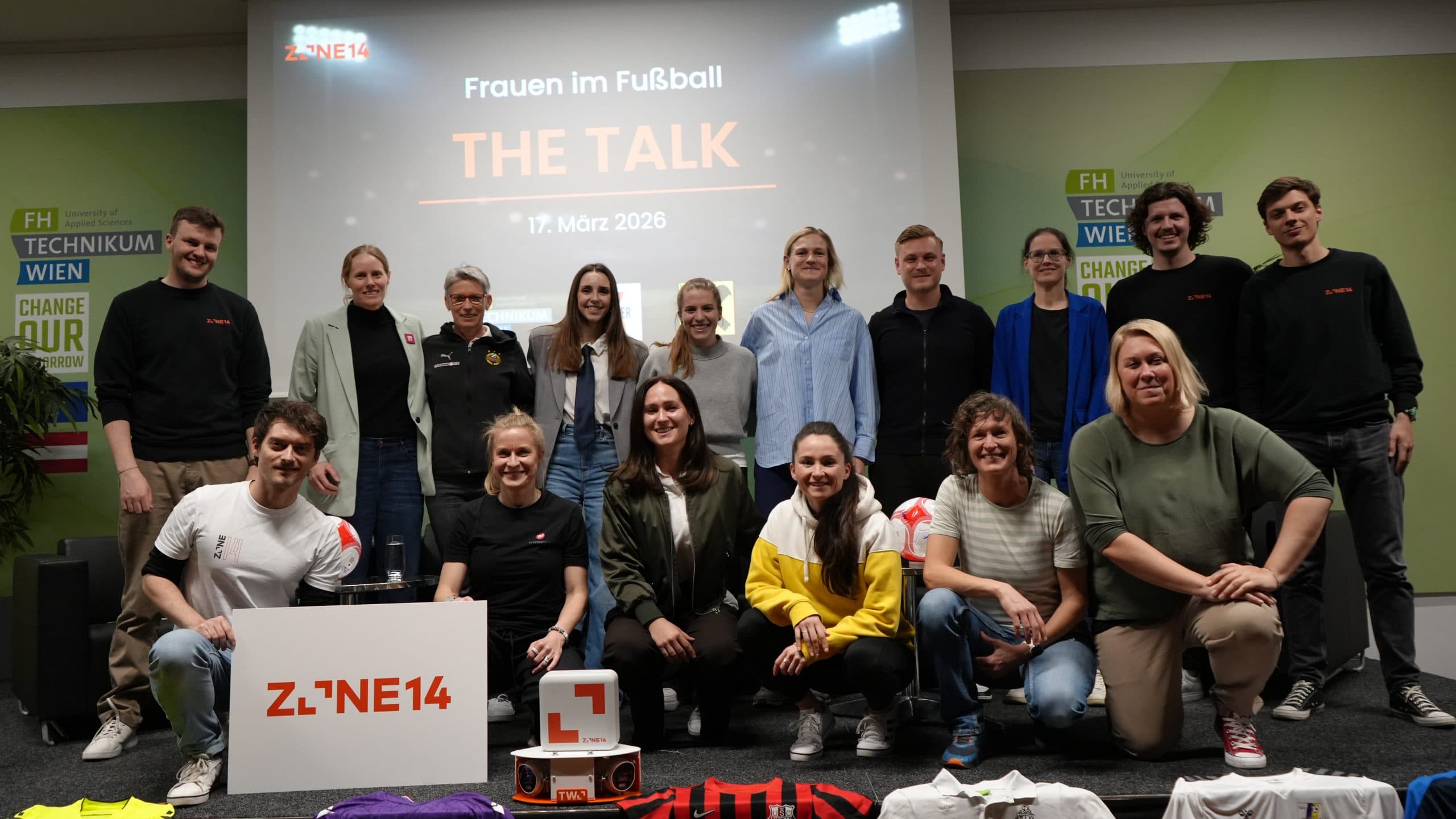 Eine Gruppe von Menschen posiert zusammen auf einer Bühne für ein Foto bei einer Veranstaltung mit dem Titel Frauen im Fußball: THE TALK am 17. März 2026. Vor ihnen sind Schilder und Fußballtrikots zu sehen.