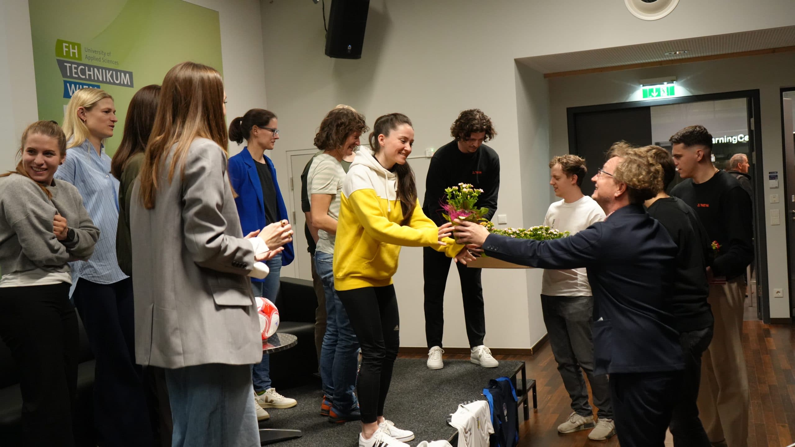Eine Gruppe von Menschen steht in einem Raum, einige lächeln, während ein Mann im Anzug einer Frau in einem gelben Kapuzenpullover Blumensträuße überreicht. Andere schauen zu und halten Blumen, und im Hintergrund ist ein Schild mit der Aufschrift TECHNIKUM zu sehen.