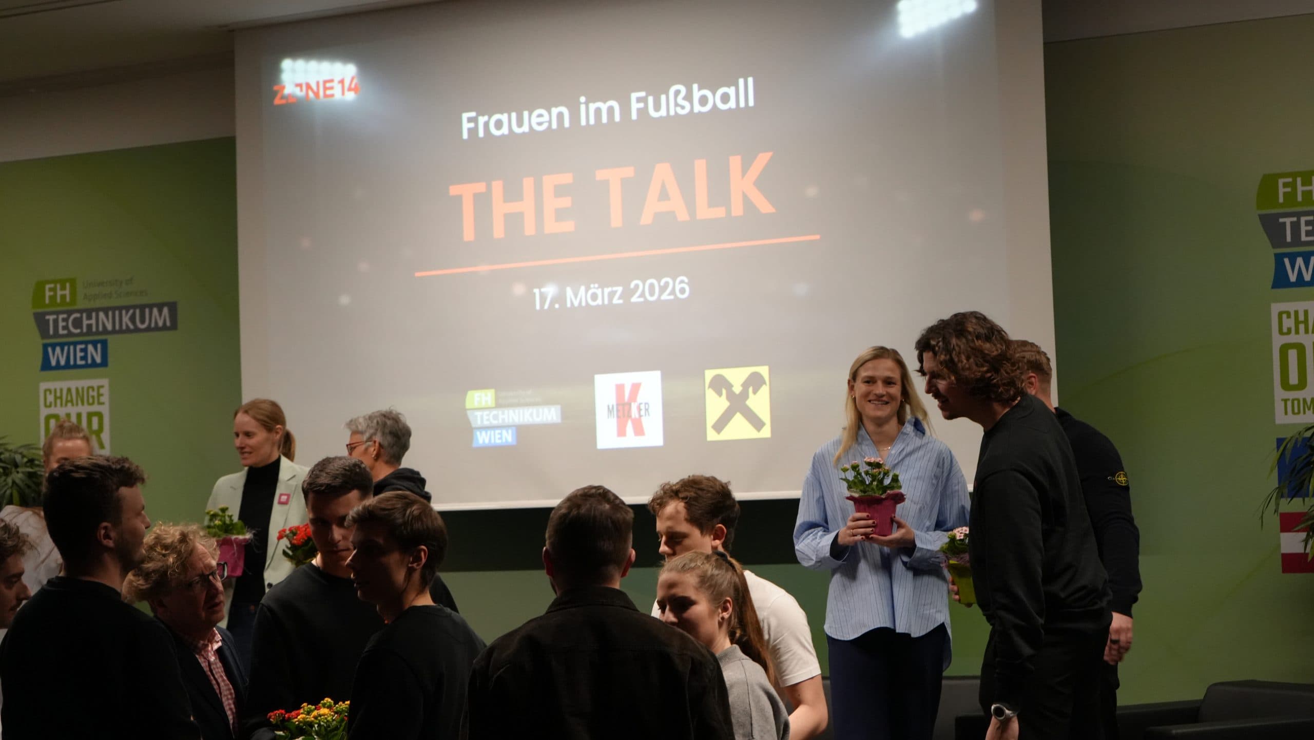 Eine Gruppe von Menschen knüpft Kontakte und hält Topfpflanzen vor einem Bildschirm, auf dem Frauen im Fußball zu sehen sind: THE TALK, 17. März 2026 bei einer Veranstaltung. Logos und Sponsorennamen sind auf der Leinwand und den Wänden zu sehen.