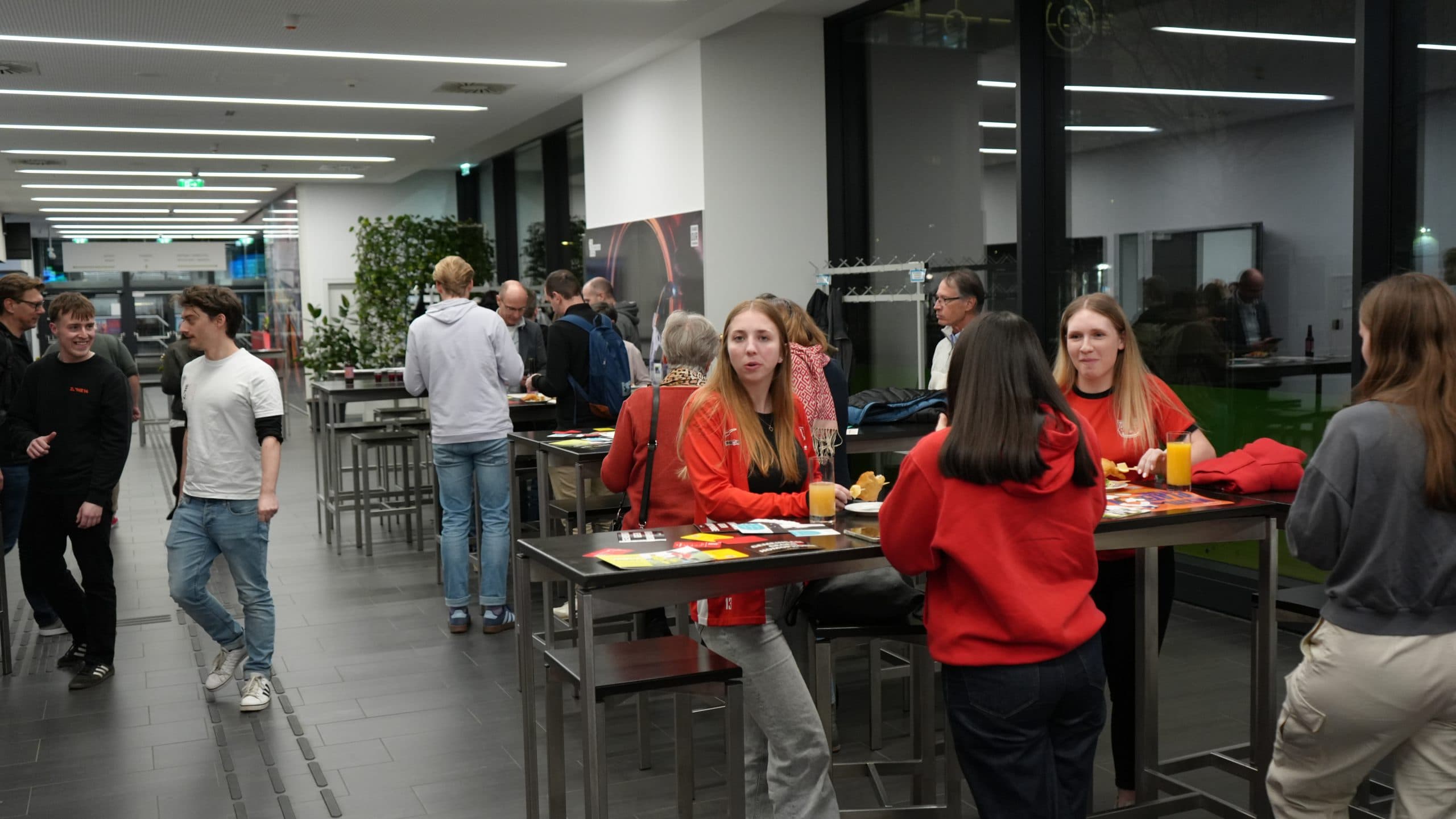 Eine Gruppe von Menschen trifft sich in einem modernen Innenraum. Mehrere Frauen in Rot sitzen und stehen um Stehtische mit Getränken und Snacks, während andere im Hintergrund herumlaufen.