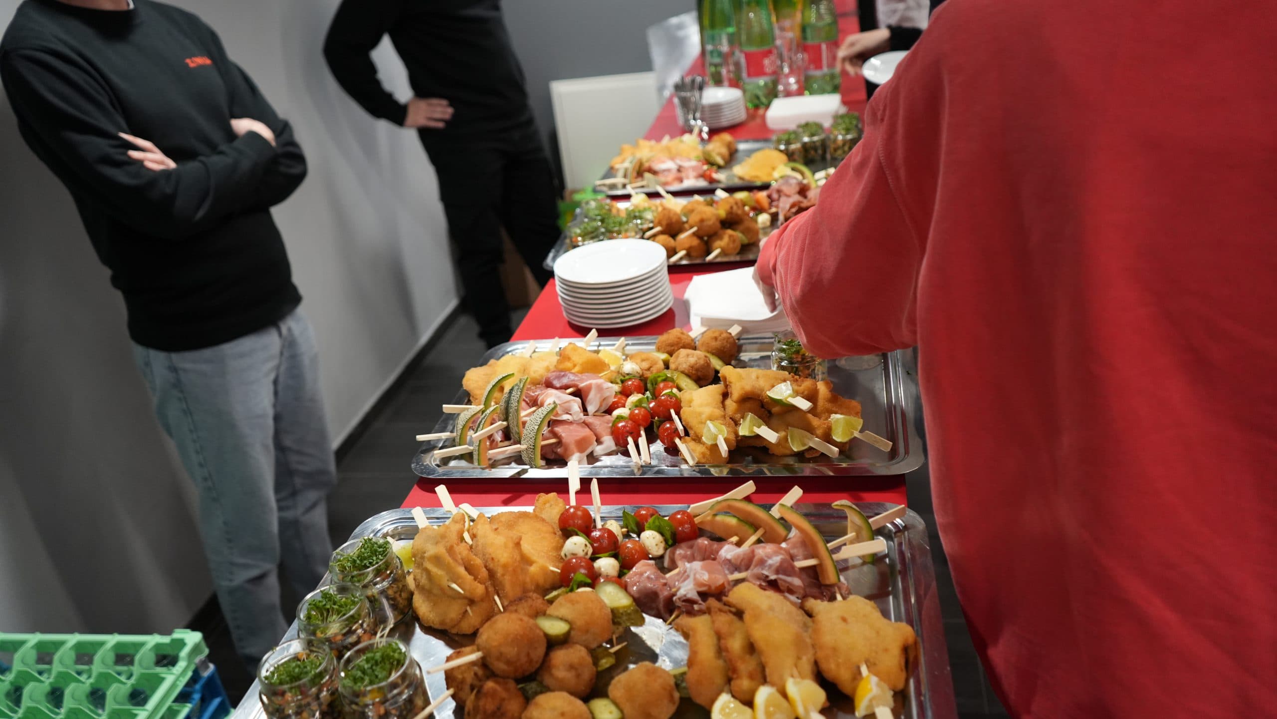 Menschen stehen um einen Tisch mit großen Tabletts mit verschiedenen Vorspeisen, darunter Spieße, frittierte Speisen und kleine Salate in Gläsern. In der Nähe sind Teller gestapelt, und eine Person bedient sich selbst von den Häppchen.