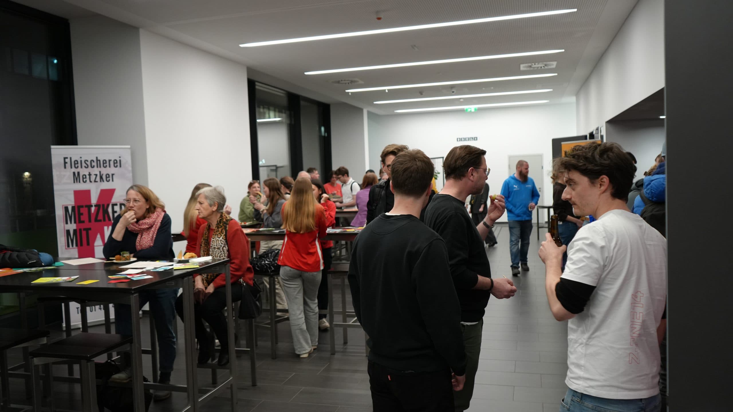 Eine Gruppe von Menschen sitzt an Stehtischen in einem hell erleuchteten Flur und isst. Im Hintergrund steht ein Schild mit der Aufschrift Fleischerei Metzker. Einige Leute sitzen, andere stehen und unterhalten sich.