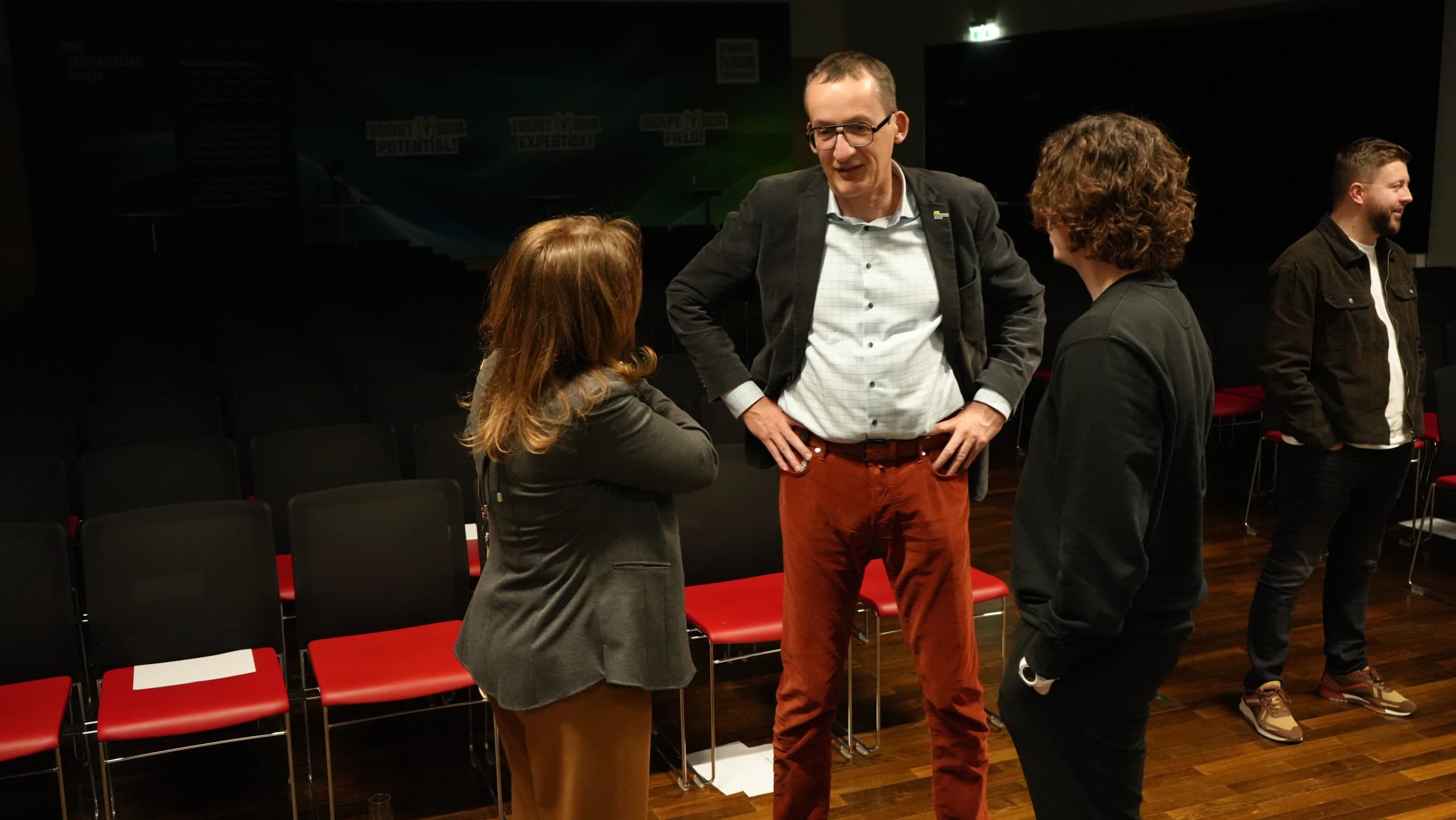 Drei Personen stehen in einem schwach beleuchteten Raum vor leeren roten und schwarzen Stühlen und unterhalten sich, während eine weitere Person allein im Hintergrund an der Wand steht.