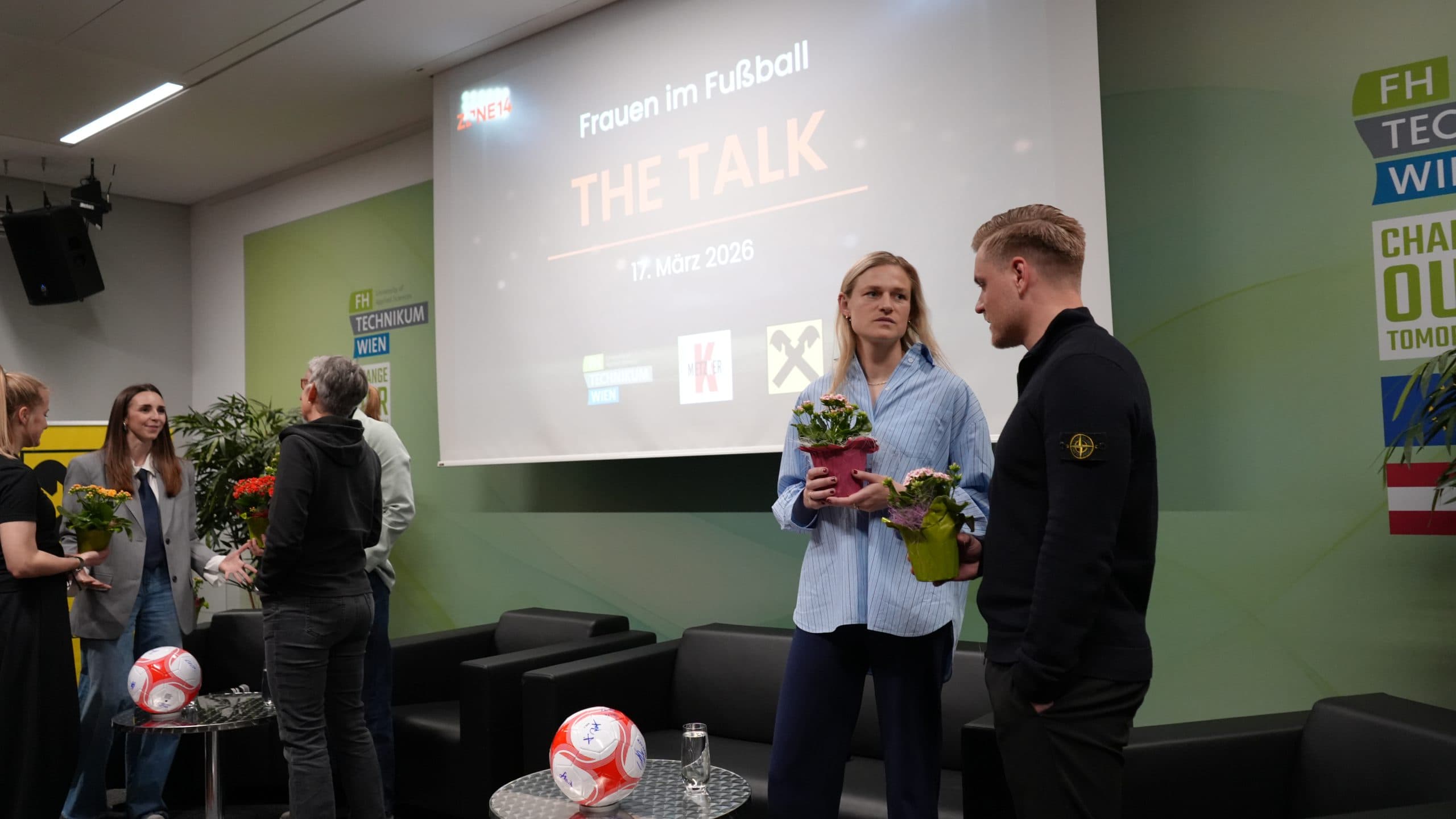 Menschen stehen und unterhalten sich vor einem Bühnenbild für eine Podiumsdiskussion mit dem Titel Frauen im Fußball: DAS GESPRÄCH. Einige halten Topfblumen in der Hand. Auf dem Tisch liegen Fußbälle und Mikrofone, als Datum ist der 17. März 2026 angegeben.