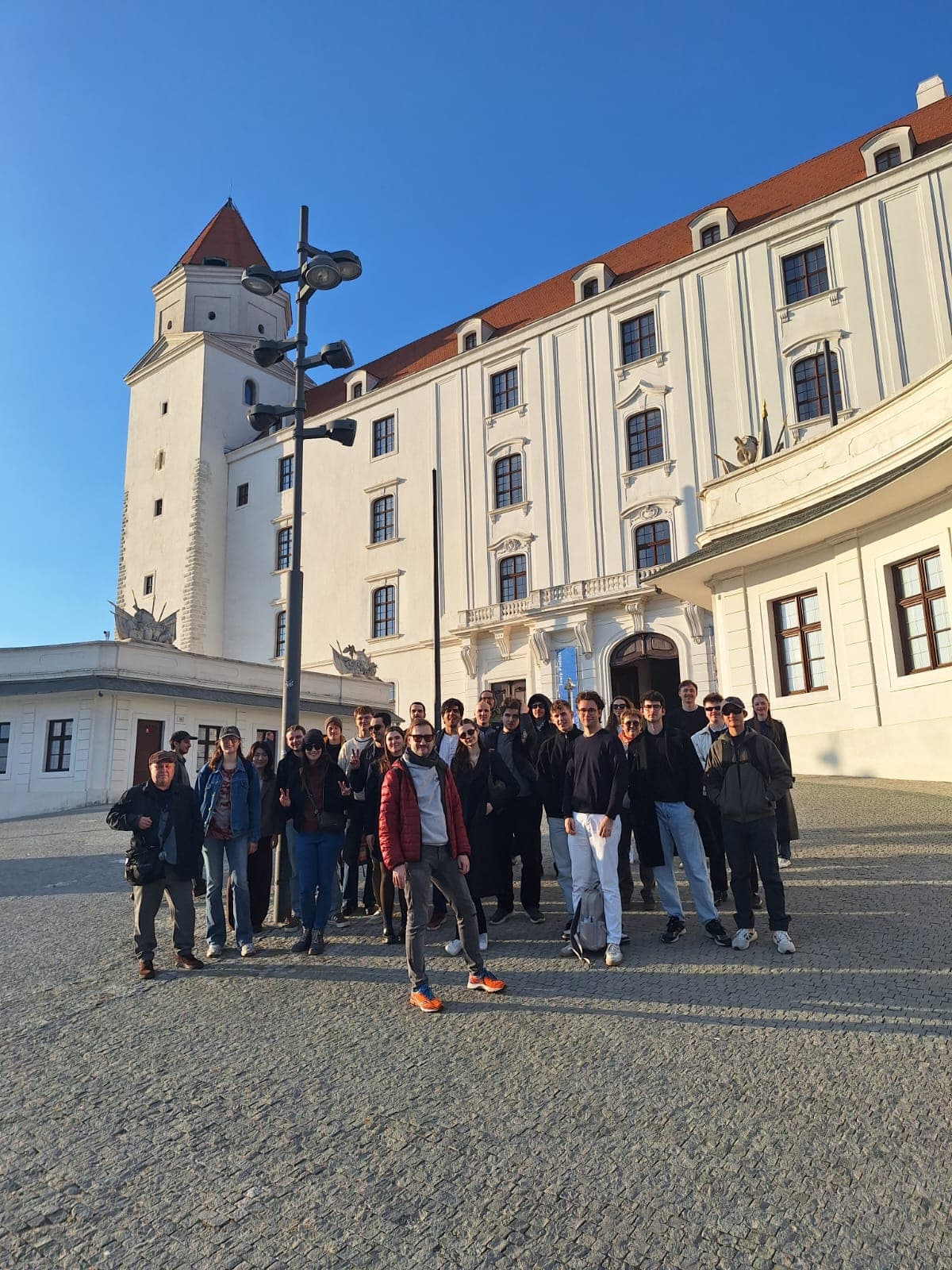 Eine Gruppe von Menschen posiert für ein Foto auf einem sonnenbeschienenen Kopfsteinpflasterplatz vor einem großen, weißen historischen Gebäude mit roten Dächern und gewölbten Fenstern. Der Himmel ist klar und blau.