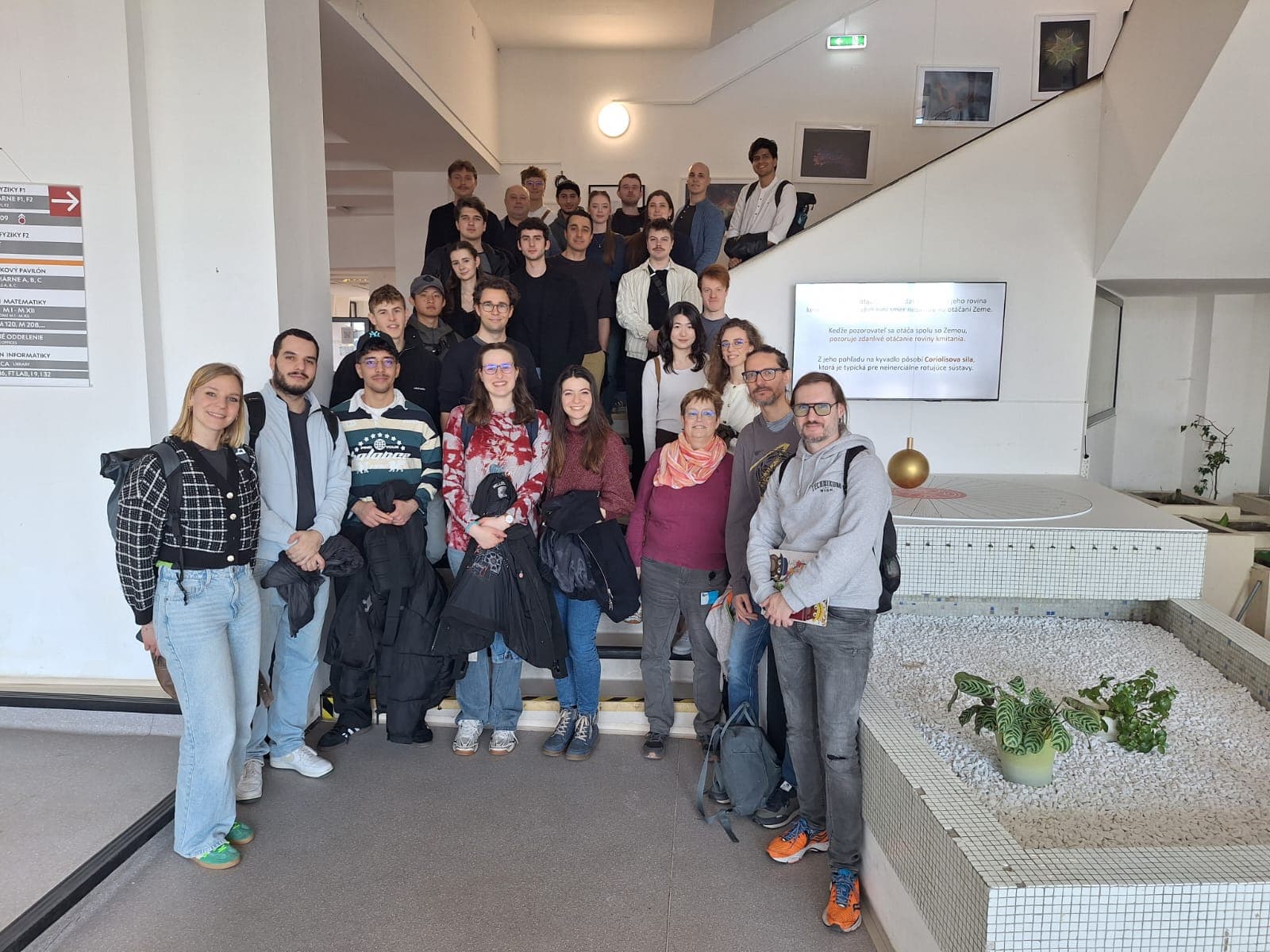 Eine Gruppe von Menschen posiert auf einer Treppe im Inneren eines modernen Gebäudes. Einige stehen auf der Treppe, andere davor. Im Vordergrund befindet sich ein weißer Übertopf mit grünen Pflanzen und einer goldenen Kugel. Alle scheinen zu lächeln.