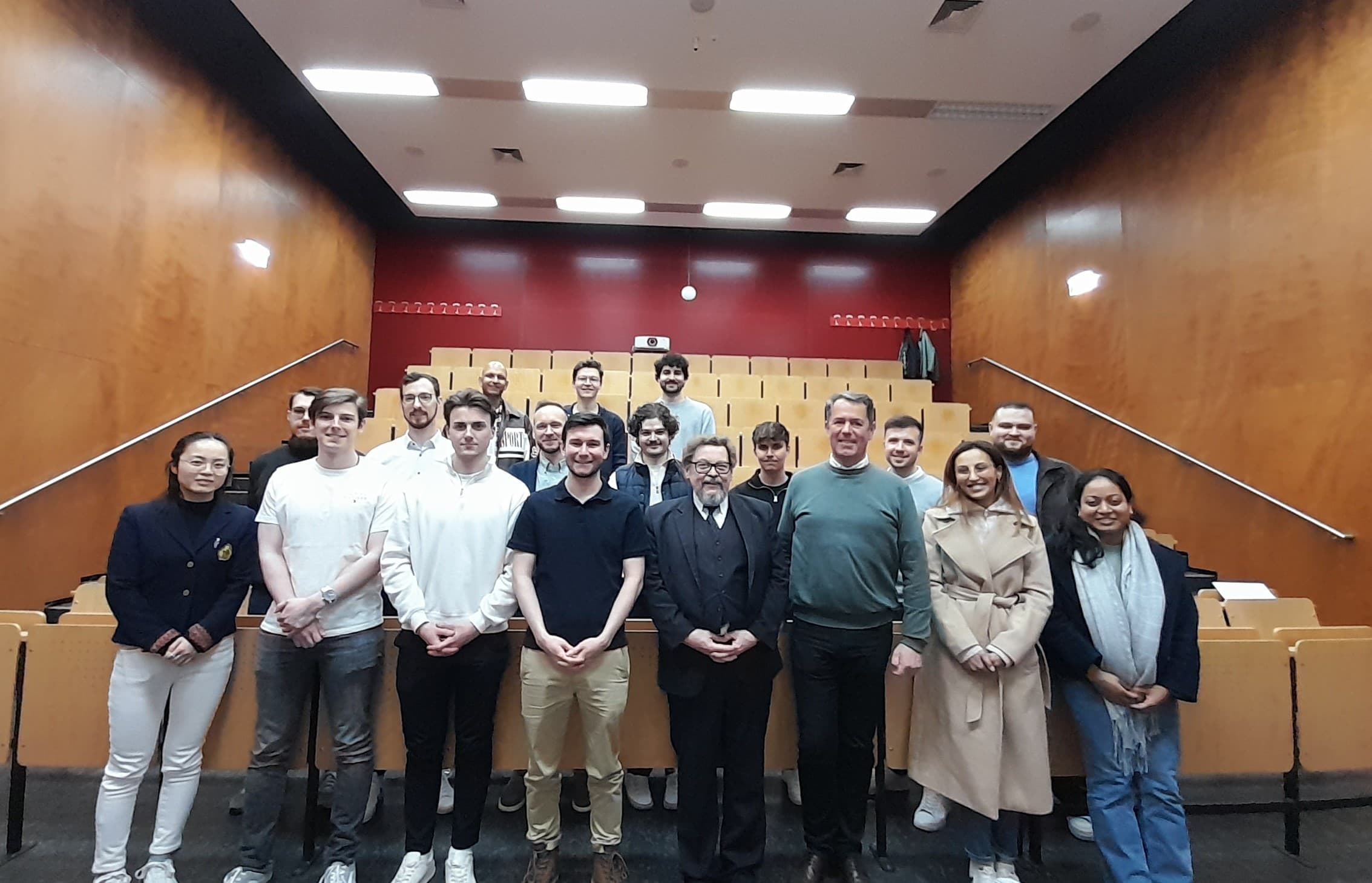 Eine Gruppe von Menschen posiert lächelnd in einem holzgetäfelten Hörsaal mit abgestufter Bestuhlung. Die meisten stehen in Reihen auf der untersten Ebene, ein paar sitzen weiter oben im Hintergrund.