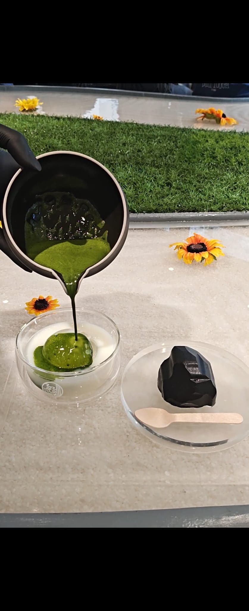 Eine Hand gießt dicke grüne Matcha-Sauce aus einem kleinen schwarzen Krug auf ein Dessert in einer Glasschüssel, daneben eine dunkle Leckerei auf einem Teller mit einem Holzlöffel. Im Hintergrund sind gelbe Blumen und ein Rasenstück zu sehen.
