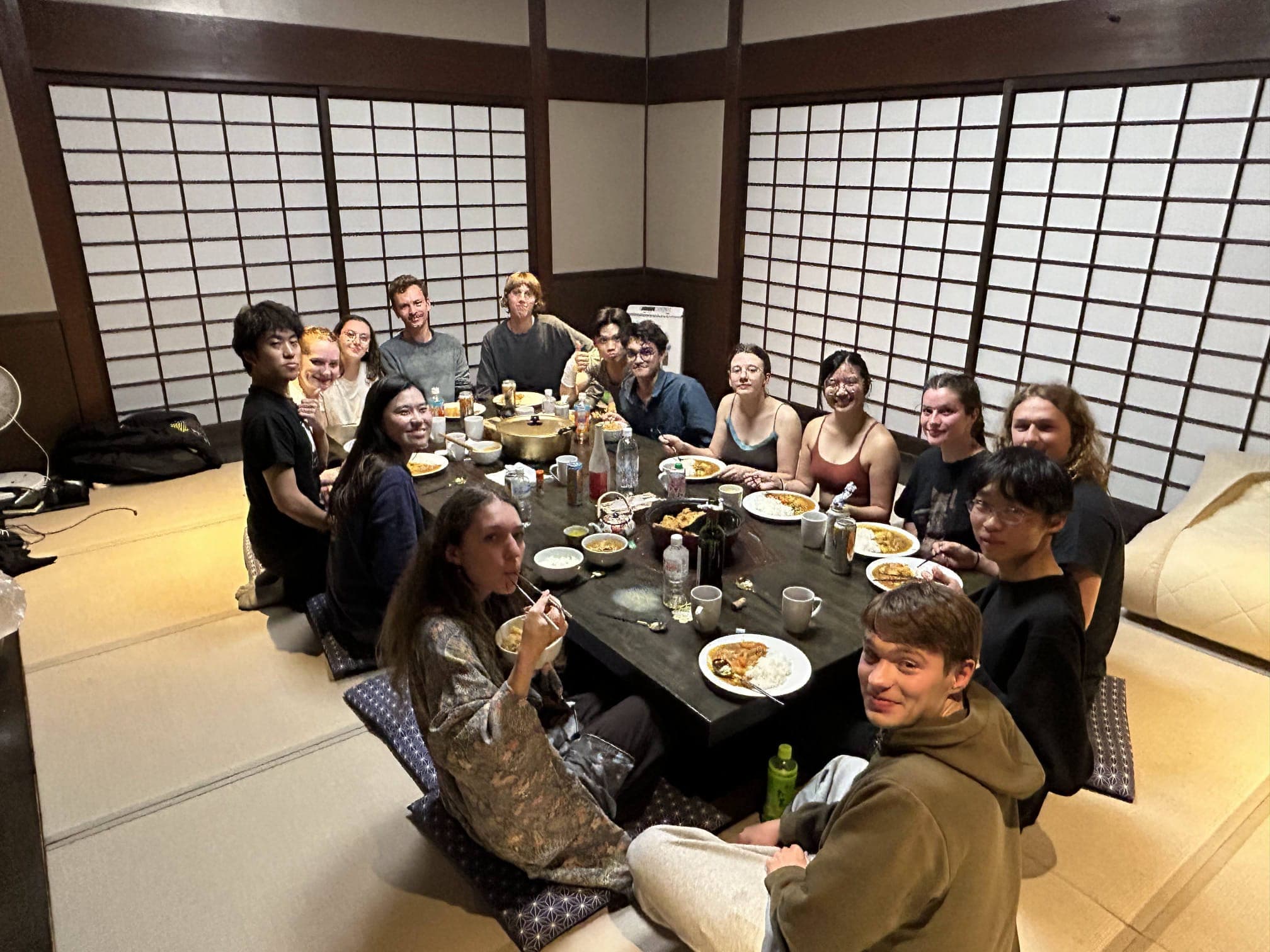 Eine Gruppe von vierzehn Personen sitzt um einen niedrigen Tisch herum und genießt eine gemeinsame Mahlzeit in einem traditionellen japanischen Raum mit Tatami-Matten und Shoji-Schirmen. Teller mit Speisen und Getränken sind auf dem Tisch verteilt.