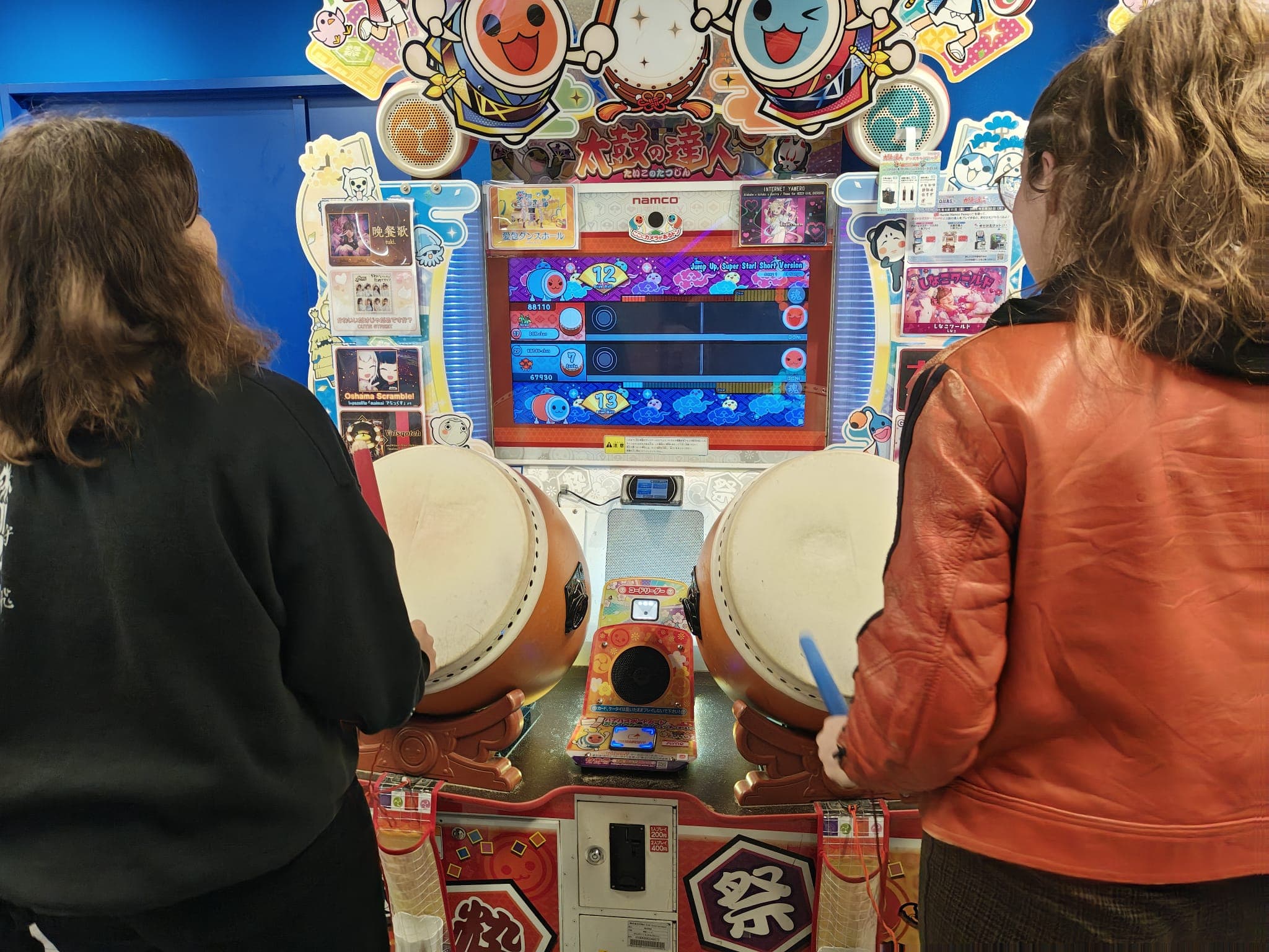 Zwei Personen spielen das Arcade-Trommelspiel Taiko no Tatsujin. Sie stehen vor dem farbenfrohen Gerät, halten beide Trommelstöcke in der Hand und bereiten sich darauf vor, auf große Taiko-Trommeln zu schlagen, während der Spielbildschirm das Spielgeschehen anzeigt.