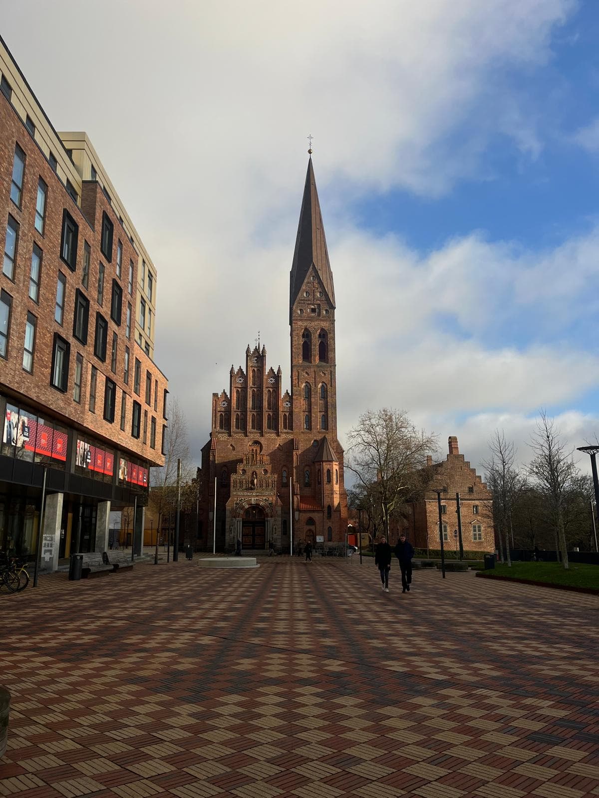 Eine hohe, spitze Kirche mit roten Backsteinmauern steht am Ende eines gemusterten Platzes, flankiert von modernen Gebäuden auf der linken Seite und Bäumen auf der rechten Seite unter einem teilweise bewölkten Himmel. Ein paar Menschen gehen auf die Kirche zu.