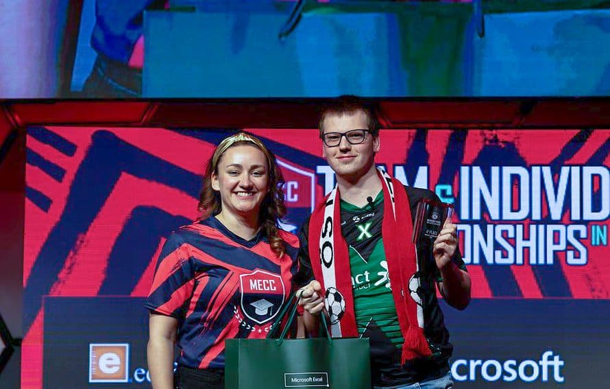 Zwei Personen, eine in einem rot-schwarzen Hemd und die andere in einem grünen Hemd mit rotem Schal, stehen auf einer esports-Veranstaltung auf der Bühne und halten Trophäen, während im Hintergrund ein Schild mit der Aufschrift Individual Championships zu sehen ist.
