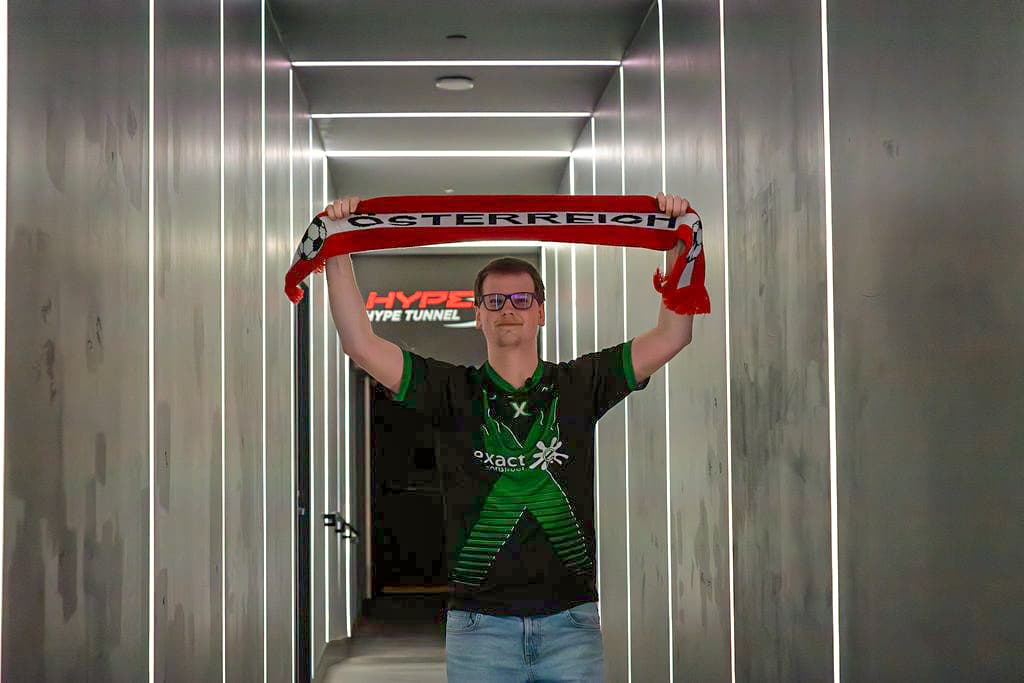 Eine Person mit Brille und einem grün-schwarzen Trikot steht in einem modernen Hausflur und hält einen rot-weißen Österreich-Schal über ihren Kopf. Im Hintergrund leuchtet das Wort HYPE auf.