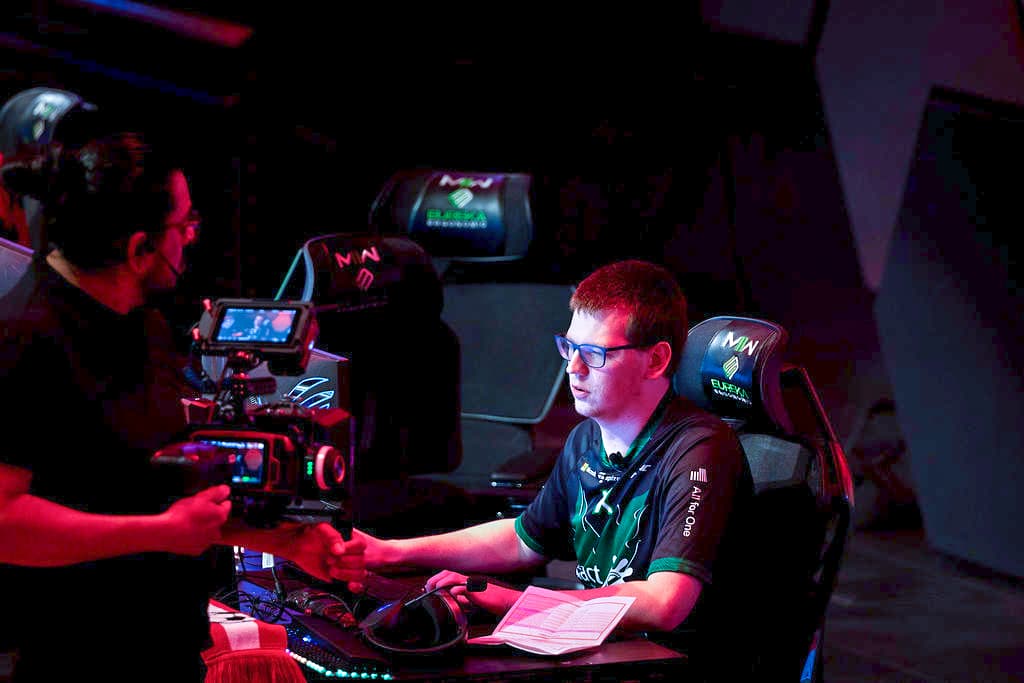 Eine Person, die eine Brille und ein Spieltrikot trägt, sitzt an einem Computertisch mit einem Headset um den Hals und schüttelt einer anderen Person, die eine Kamera hält, die Hand. Spielesessel und elektronische Geräte sind zu sehen.