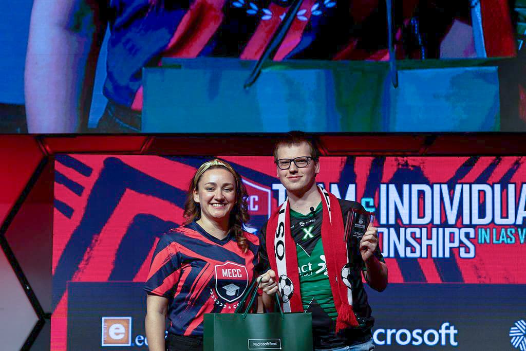 Zwei esports-Spieler stehen auf der Bühne, halten Trophäen und lächeln. Sie tragen Mannschaftstrikots und Schals, hinter ihnen hängt ein rot-schwarzes Meisterschaftsbanner.