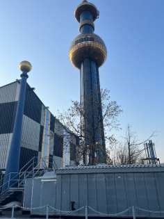 Ein hohes, zylindrisches Gebäude mit goldenen Akzenten und einer runden Aussichtsplattform steht neben einem gestreiften Industriegebäude unter einem klaren blauen Himmel. Im Vordergrund sind Bäume und ein kleiner Zaun zu sehen.