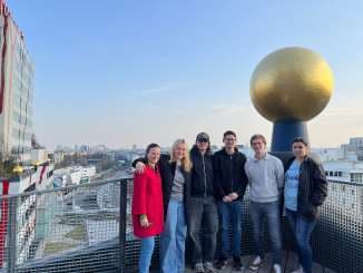 Sechs Personen stehen zusammen auf einem Dach mit einer Stadtlandschaft im Hintergrund. Sie lächeln, und eine große goldene Kugel auf einem Sockel steht hinter ihnen. Der Himmel ist klar und blau.