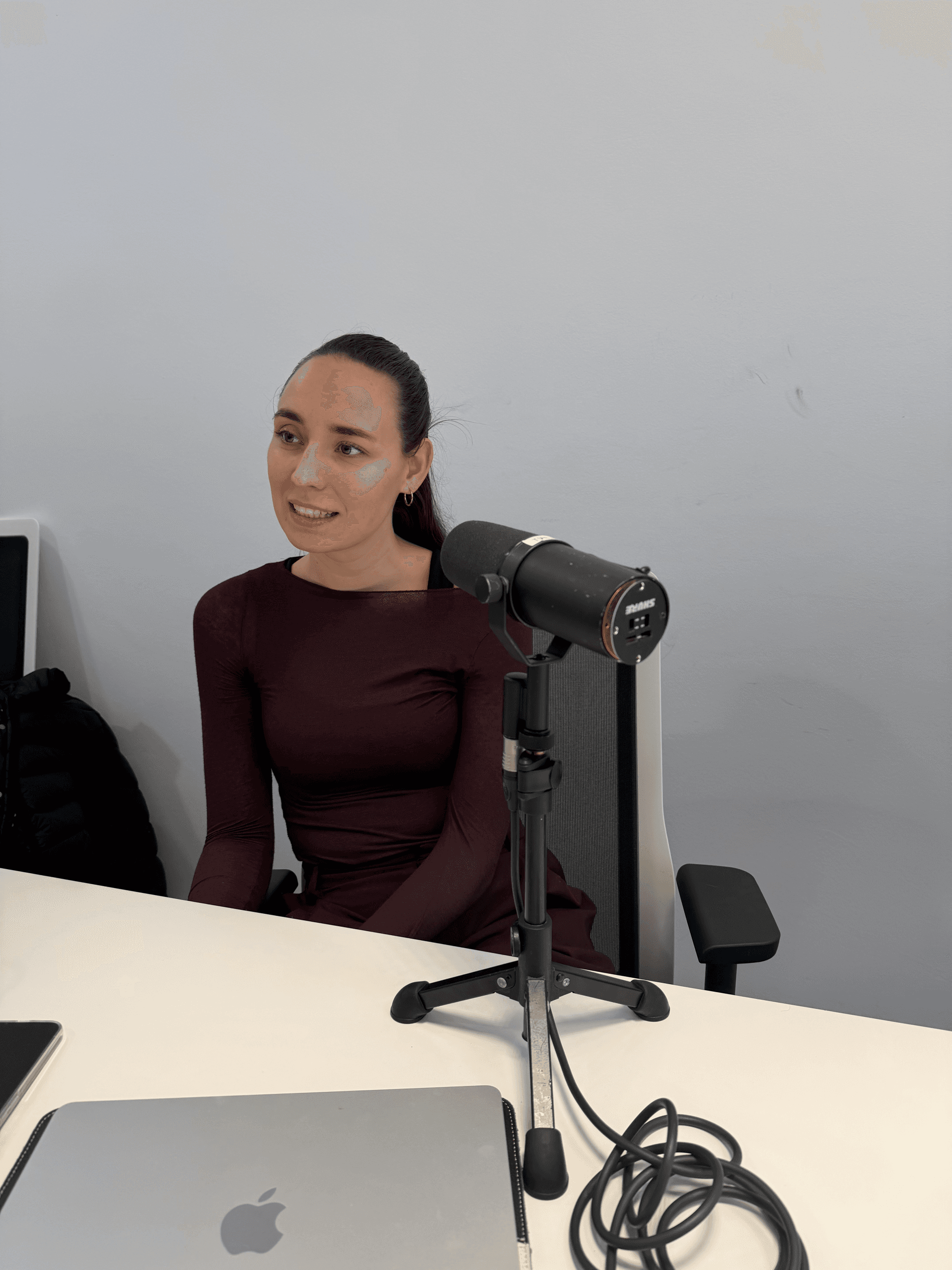 Eine Frau in einem kastanienbraunen Kleid sitzt an einem Schreibtisch mit einem Mikrofon auf einem Ständer, einem Laptop und Kabeln. Sie scheint in einer modernen, minimalistischen Büroumgebung zu sprechen oder aufzunehmen.