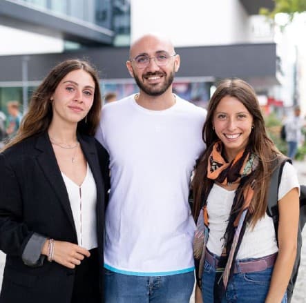 Drei Personen stehen im Freien dicht beieinander und lächeln in die Kamera. Der Mann in der Mitte trägt eine Brille und ein weißes Hemd, während die beiden Frauen zu beiden Seiten leger gekleidet sind, eine mit einem Schal, die andere mit einer schwarzen Jacke.