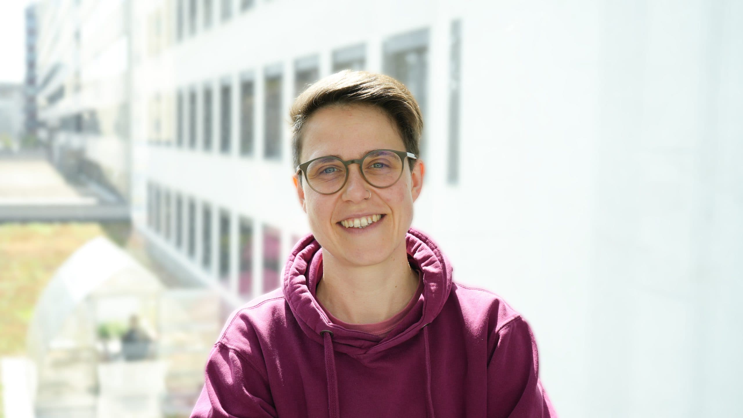 Eine Person, die eine Brille und einen kastanienbraunen Kapuzenpullover trägt, lächelt in die Kamera, während sie in einem Innenraum steht, in dem helles natürliches Licht und ein verschwommenes Gebäude im Hintergrund zu sehen sind.