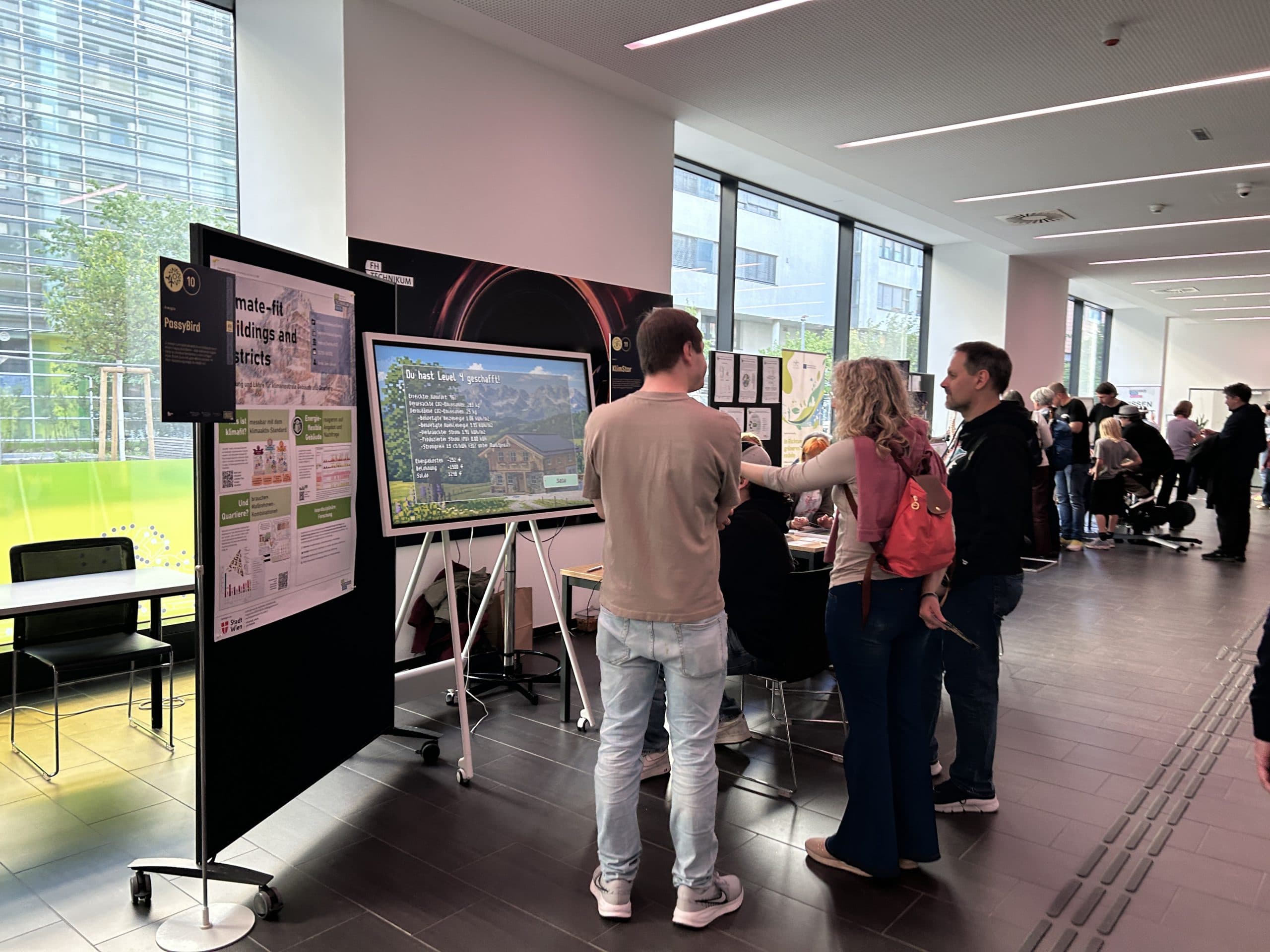 Menschen stehen und sprechen in einer Indoor-Wissenschafts- oder Technologieausstellung mit Postern und einem großen Monitor, der Informationen anzeigt. Die Veranstaltung findet in einer modernen, gut beleuchteten Halle mit großen Fenstern statt.