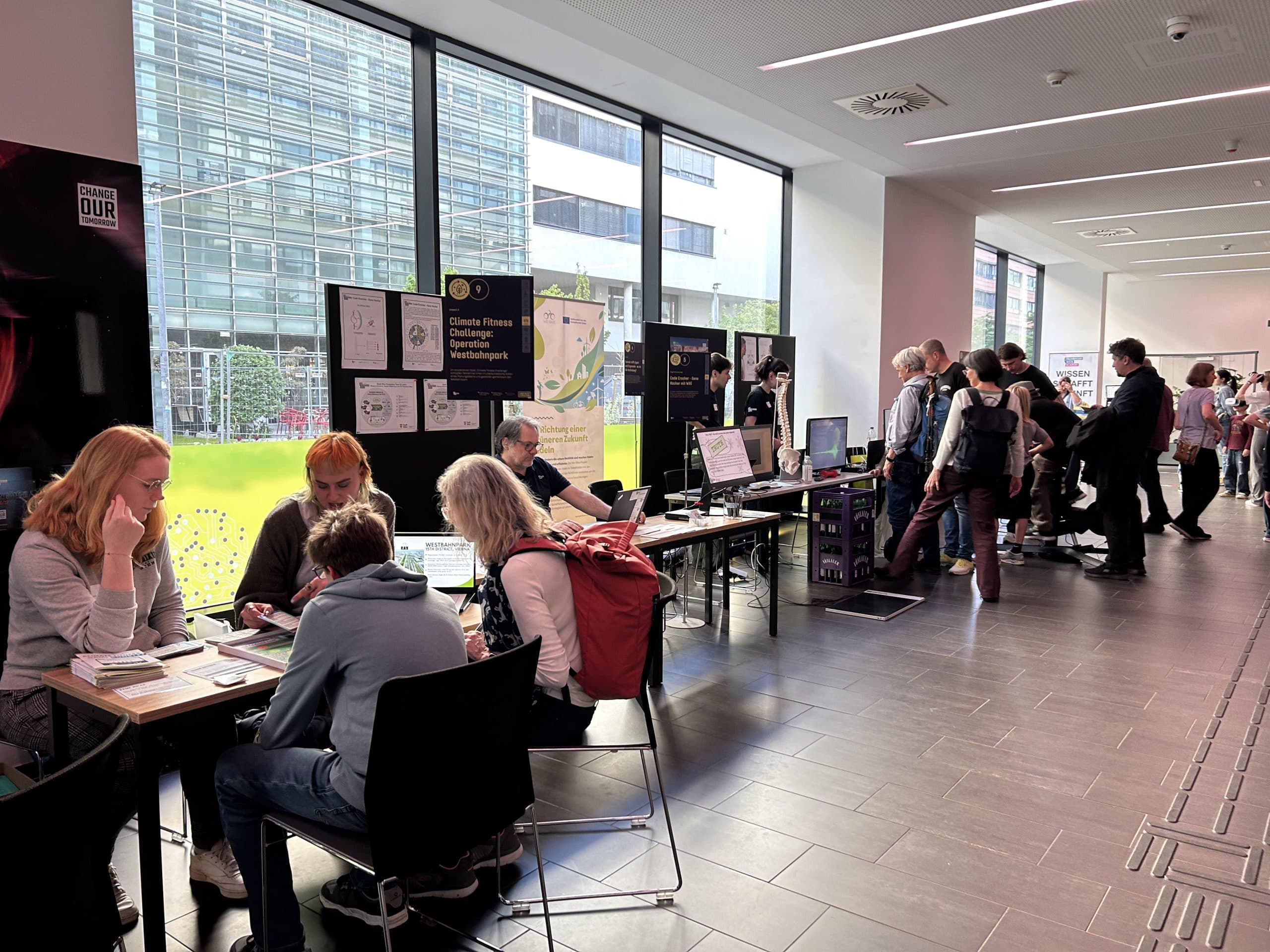 Bei einer Indoor-Veranstaltung mit großen Fenstern besuchen die Besucher die Stände; einige sitzen an Tischen mit Broschüren und Laptops, während andere stehen und mit den Ausstellern ins Gespräch kommen. Hinter den Tischen sind Plakate und Displays zu sehen.