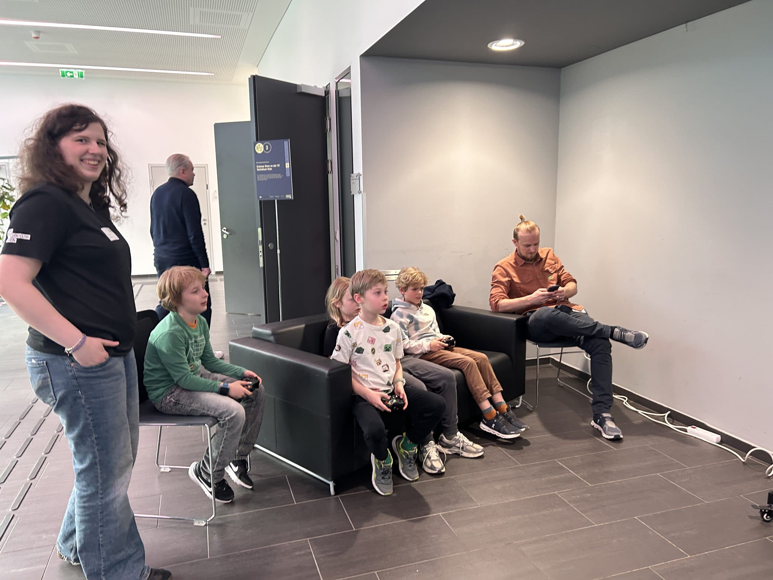 Mehrere Kinder sitzen auf Stühlen und spielen Videospiele mit Controllern, während ein Erwachsener in der Nähe sitzt und ein Tablet benutzt. Ein weiterer Erwachsener steht lächelnd im Hintergrund. Der Schauplatz scheint ein öffentlicher Raum oder ein Veranstaltungsort zu sein.