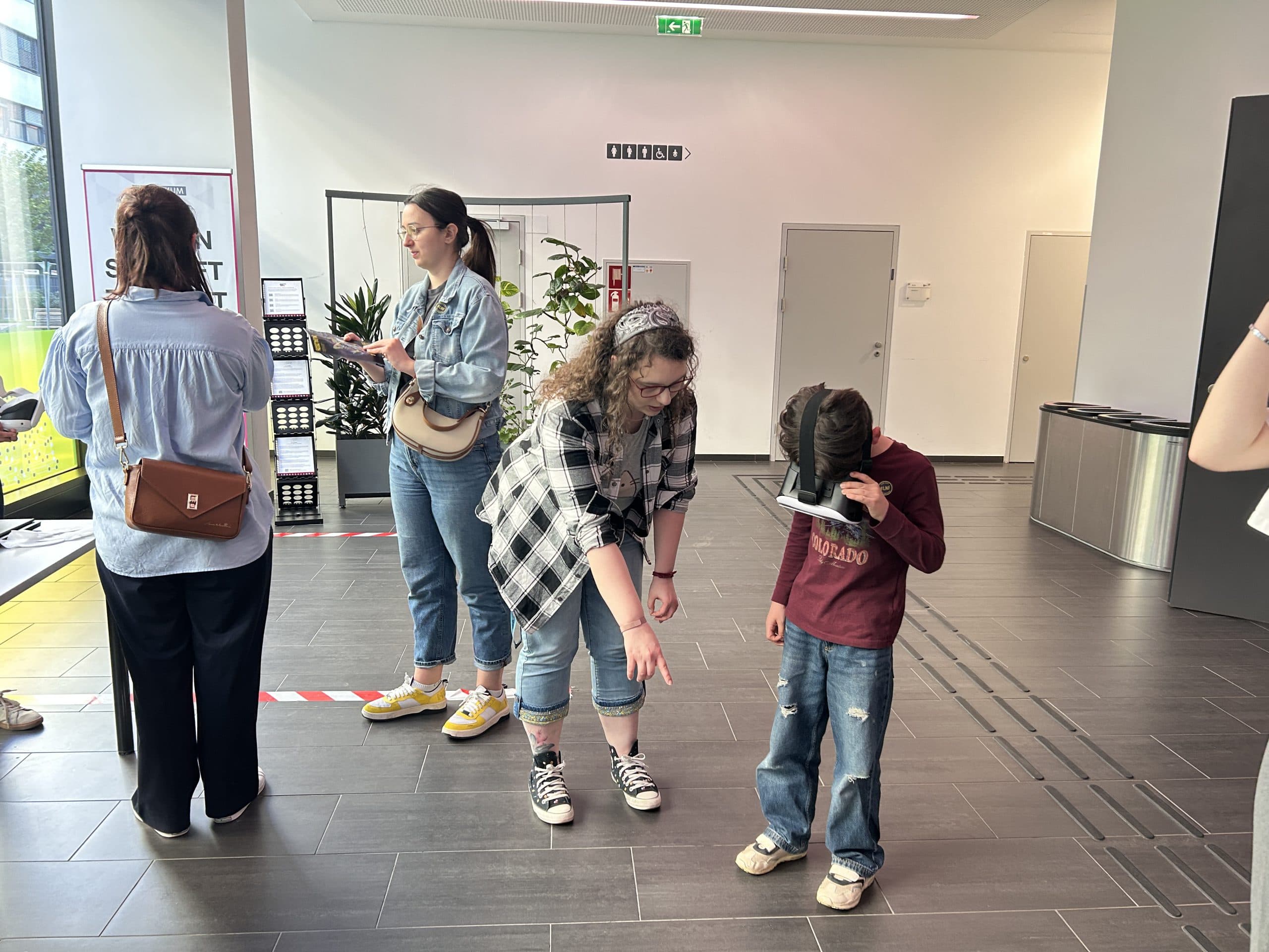 Eine Frau in einem karierten Hemd zeigt auf den Boden, während ein Kind neben ihr auf ein Tablet schaut. Zwei weitere Personen stehen in der Nähe, eine von ihnen benutzt ein Smartphone. Die Szene spielt sich in einer modernen Eingangshalle ab.