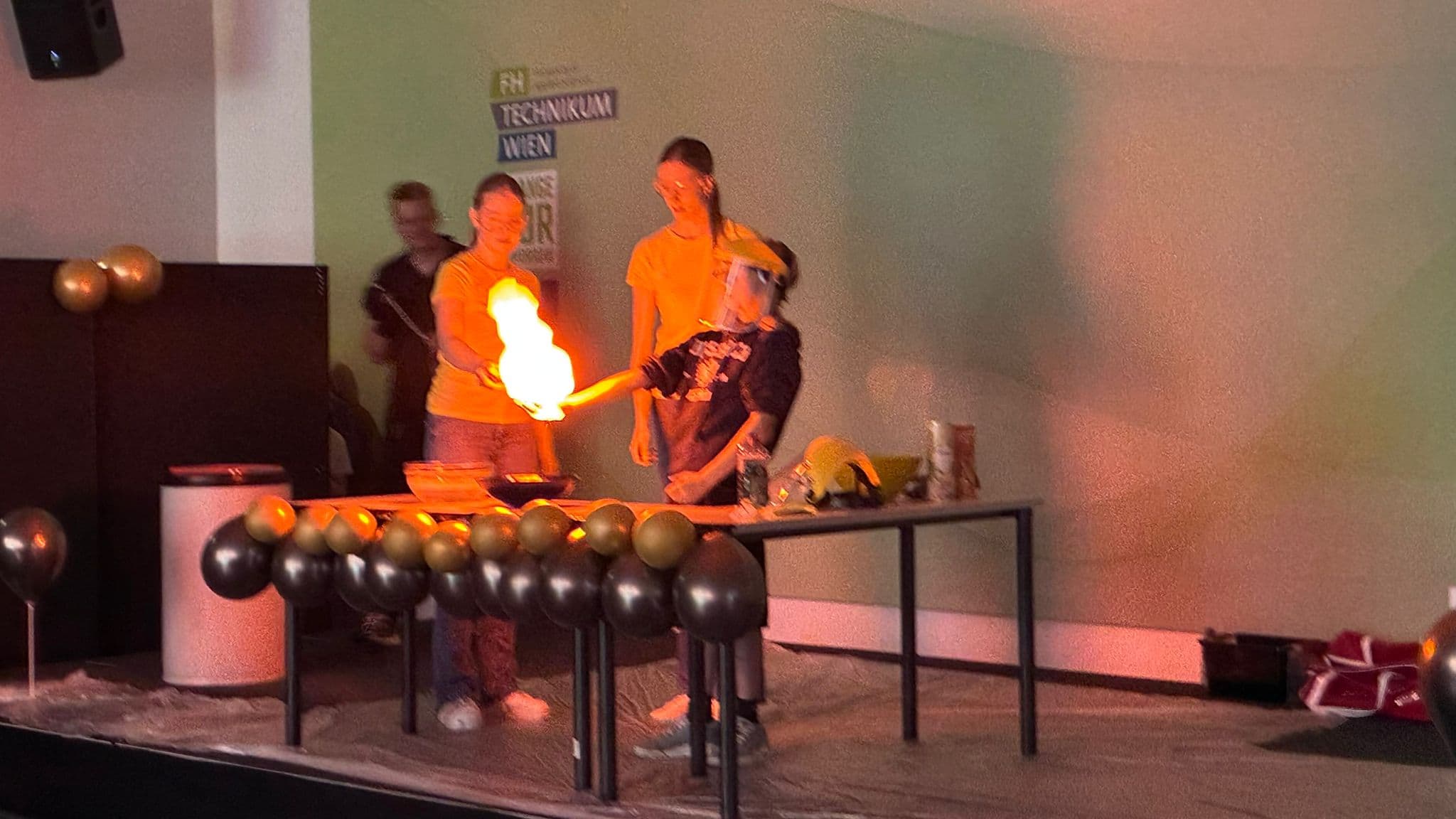 Ein Kind, das von zwei Erwachsenen unterstützt wird, führt auf der Bühne ein wissenschaftliches Experiment durch, bei dem es einen Feuerstoß aus seiner Hand erzeugt. Die Szene ist mit schwarzen und goldenen Luftballons dekoriert.