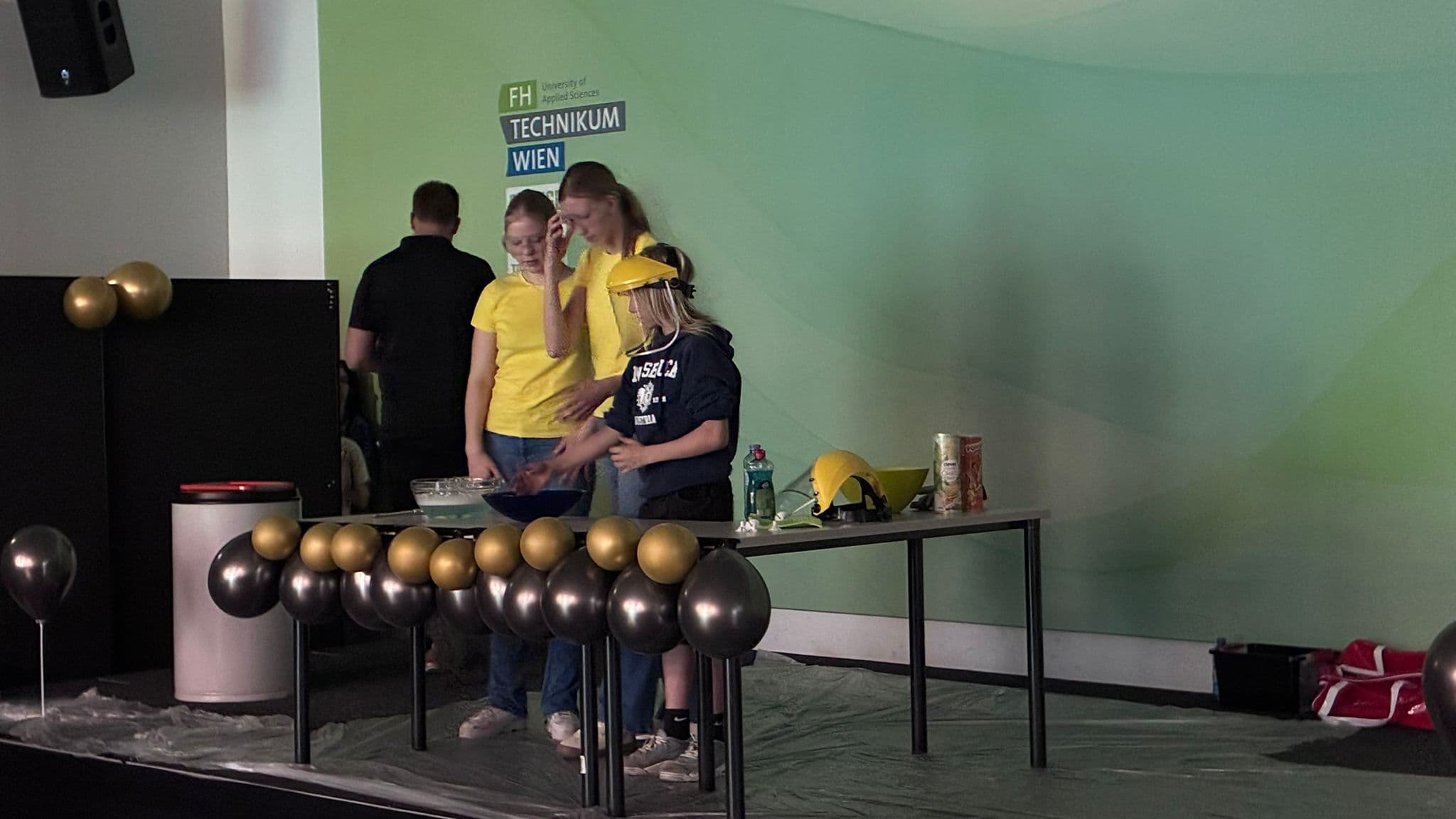 Drei Personen mit gelben Hemden und Hüten stehen hinter einem Tisch mit Snacks und Schüsseln. Im Hintergrund ist ein Schild der FH Technikum Wien zu sehen, und schwarze und goldene Luftballons schmücken die Bühne.