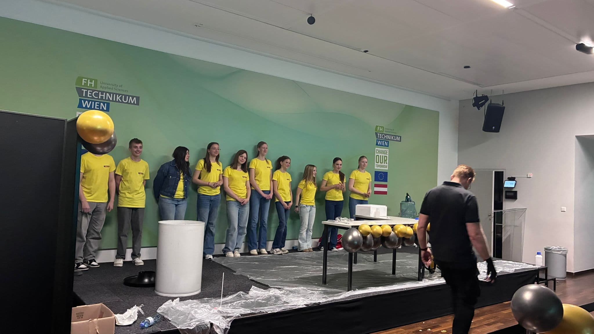 Eine Gruppe von Menschen in gelben Hemden steht auf einer Bühne an der FH Technikum Wien. Einige unterhalten sich, auf einem Tisch sind wissenschaftliche Geräte und schwarz-goldene Luftballons aufgebaut. Eine Person geht im Vordergrund.