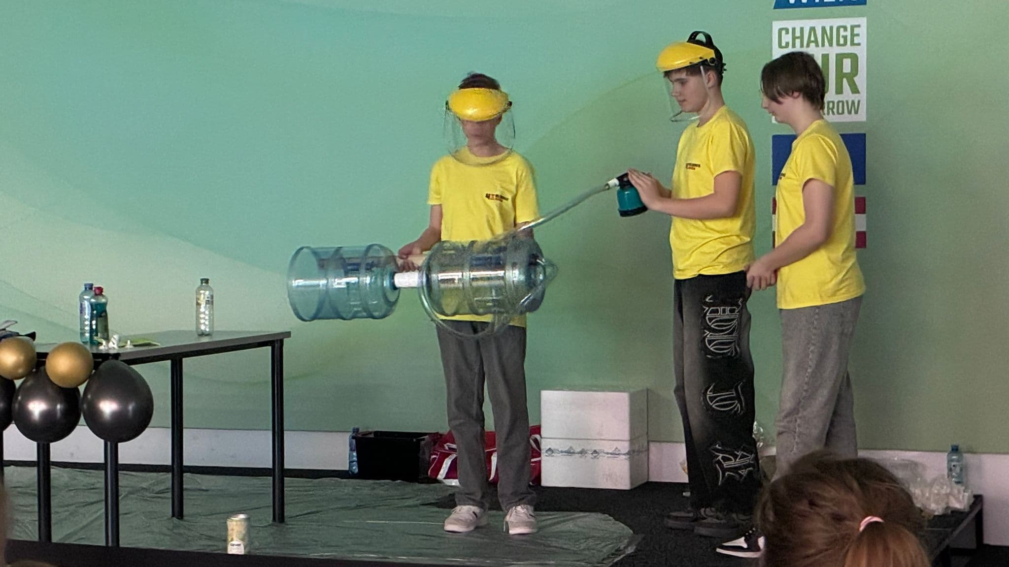 Drei junge Leute in gelben Hemden und mit Gesichtsschutz nehmen an einer wissenschaftlichen Demonstration teil, bei der große Plastikbehälter und eine Nebelmaschine auf einer Bühne mit grünen Wänden und Luftballons in der Nähe zum Einsatz kommen.