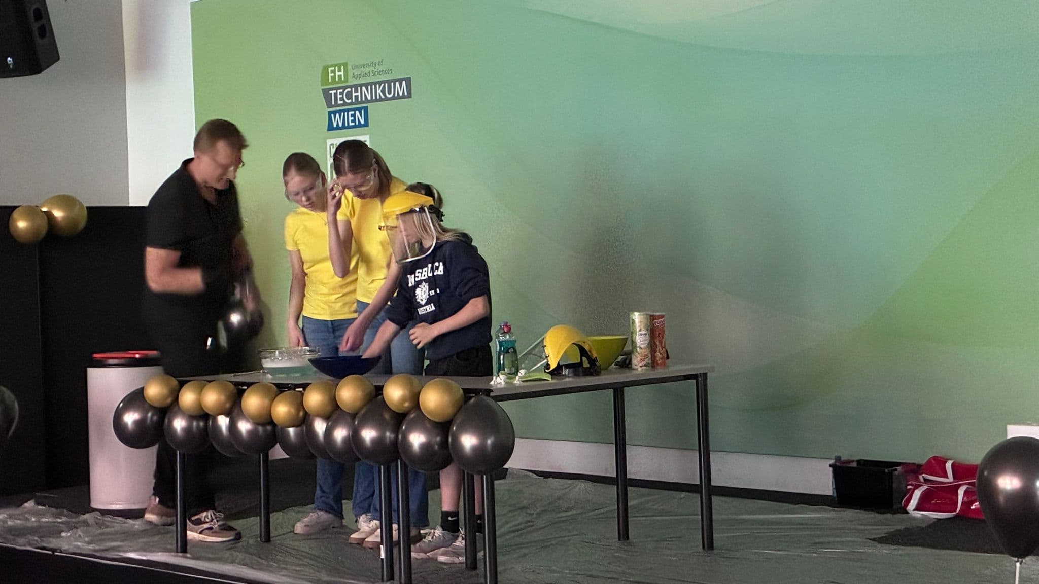 Vier Personen, drei in gelben und eine in schwarzen Hemden, stehen um einen Tisch, auf dem verschiedene Gegenstände liegen. Schwarze und goldene Luftballons schmücken die Front. Dahinter steht eine grüne Wand mit der Aufschrift FH TECHNIKUM WIEN.