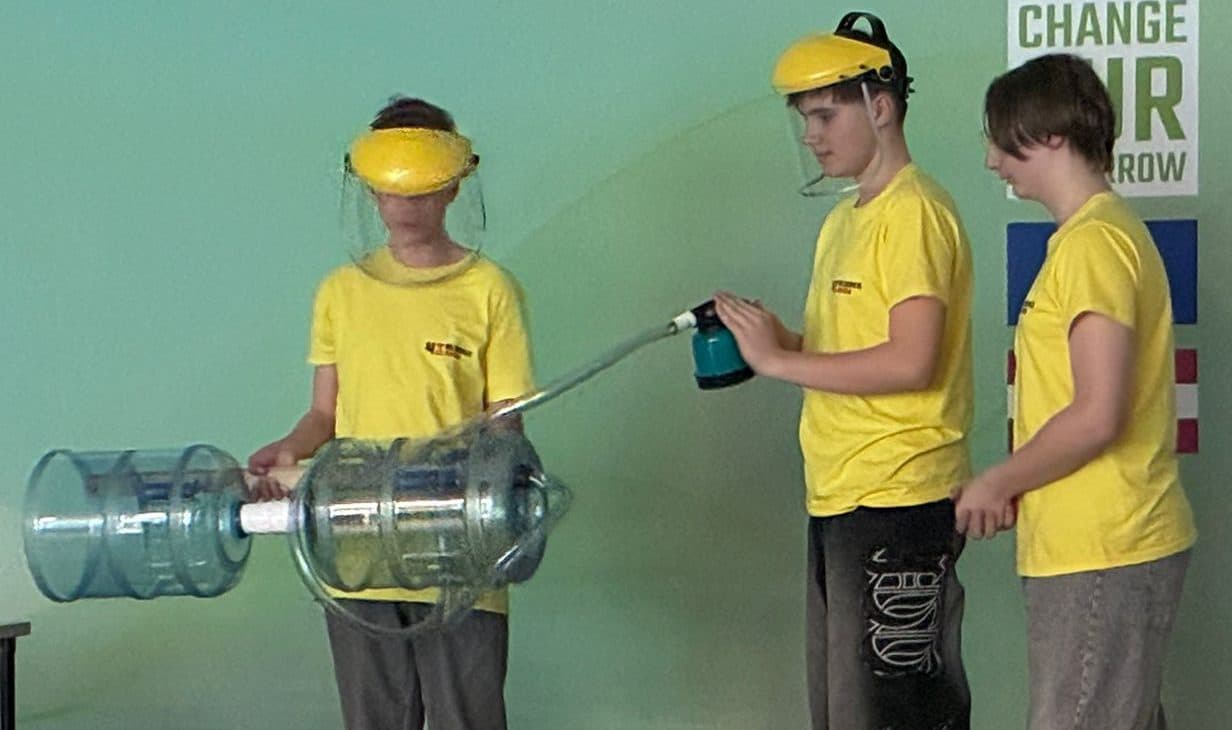 Drei junge Leute in gelben Hemden und mit Schutzvisier führen auf der Bühne ein wissenschaftliches Experiment durch. Einer hält große Plastikröhrchen, ein anderer gießt Flüssigkeit ein und der dritte beobachtet. Schwarze und goldene Luftballons schmücken die Szene.