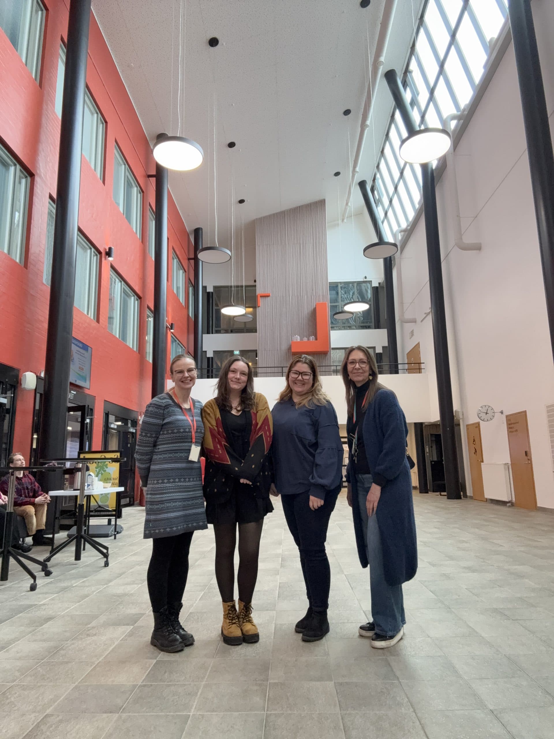 Vier Frauen stehen lächelnd in einem hellen, geräumigen Atrium mit hohen Fenstern, roten und weißen Wänden und moderner Beleuchtung. Sie posieren zusammen auf einem gefliesten Boden.