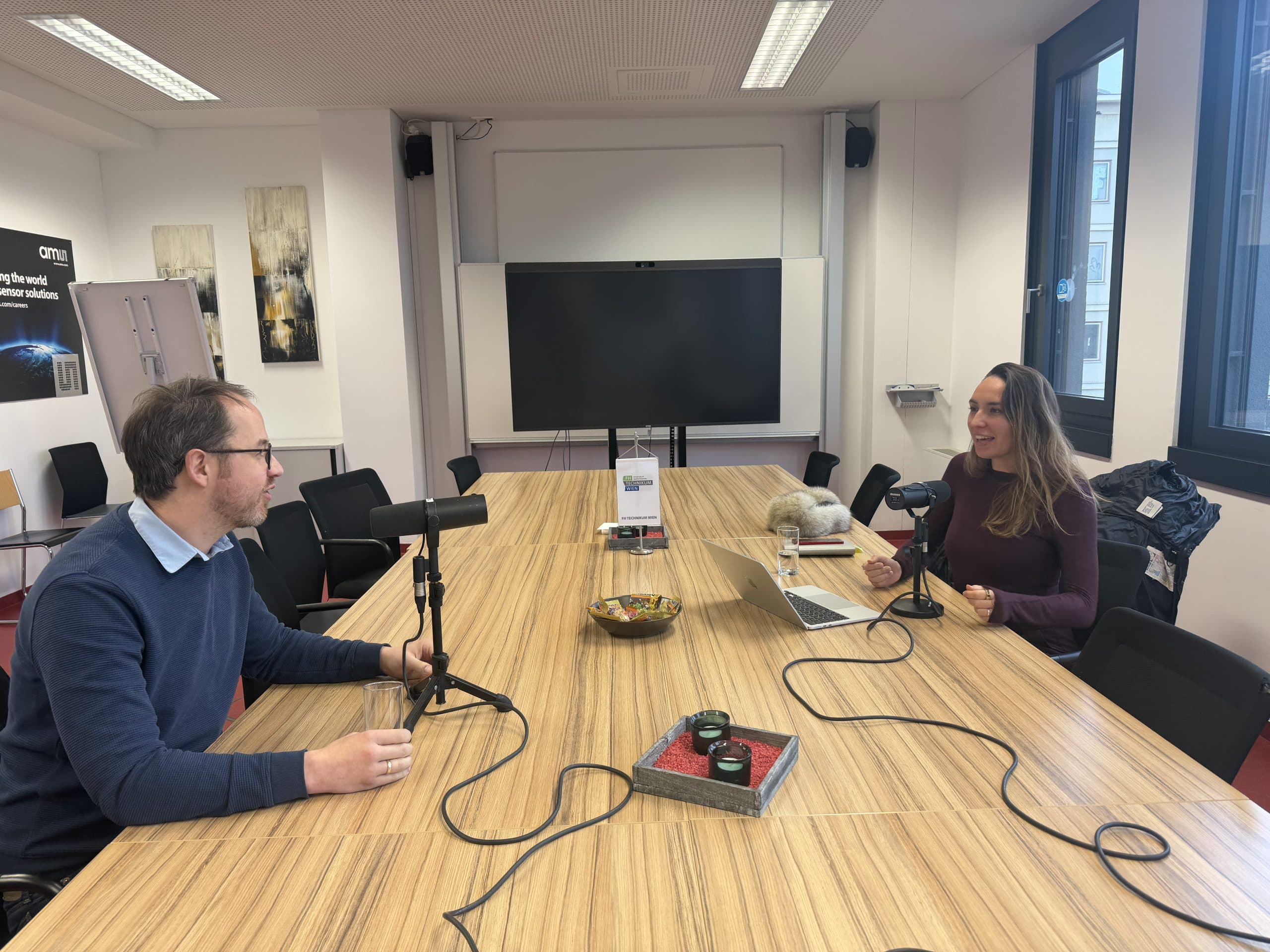 In einem modernen Konferenzraum mit großen Fenstern und einem leeren Bildschirm im Hintergrund sitzen sich zwei Personen an einem langen Holztisch mit Mikrofonen, Aufnahmegeräten und einem Laptop gegenüber.