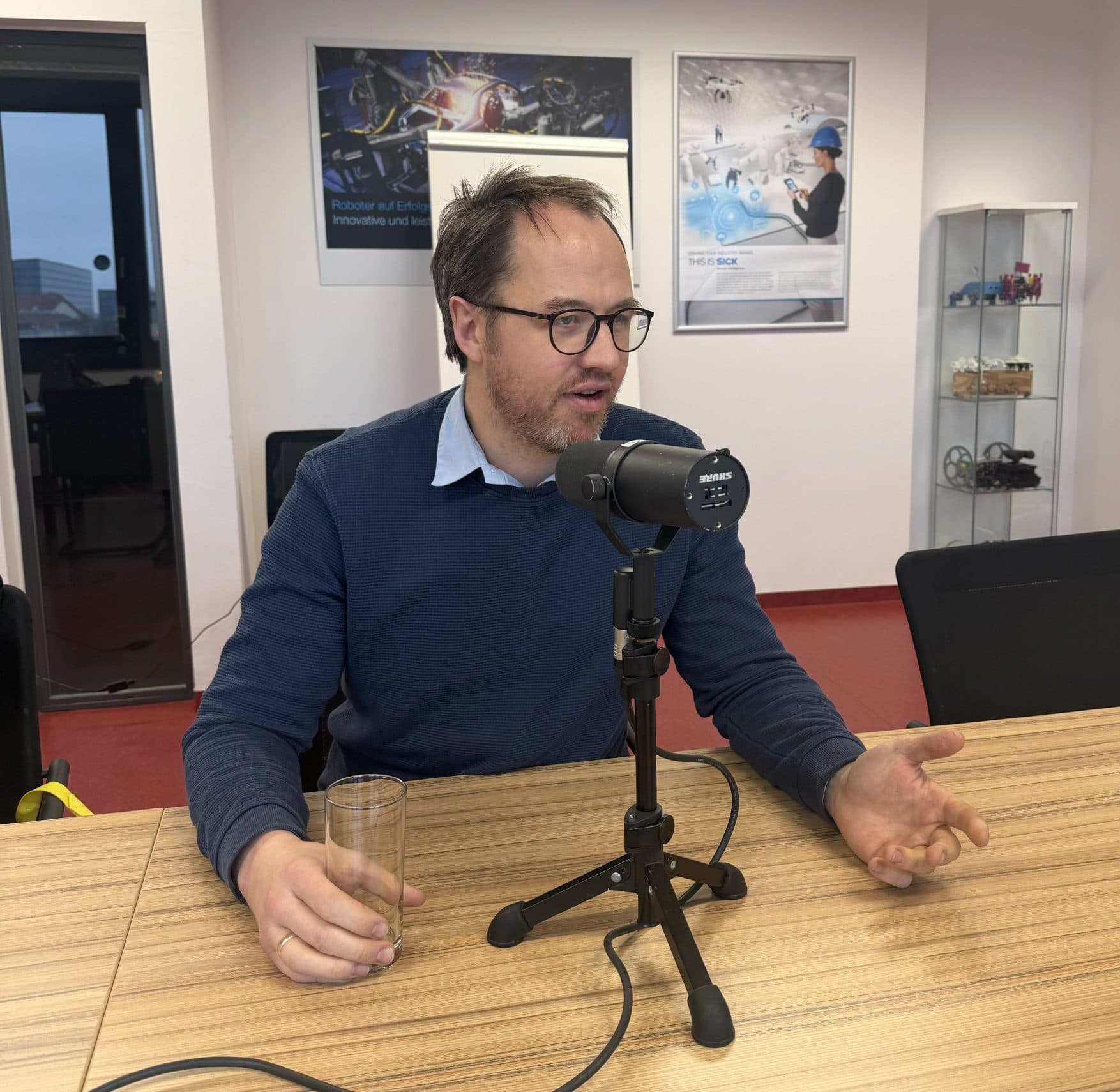 Ein Mann sitzt an einem Tisch und spricht in ein Mikrofon, in der einen Hand hält er ein Glas. Er trägt eine Brille und einen blauen Pullover. Im Hintergrund sind Poster und Regale zu sehen, die auf ein Büro oder ein Studio schließen lassen.