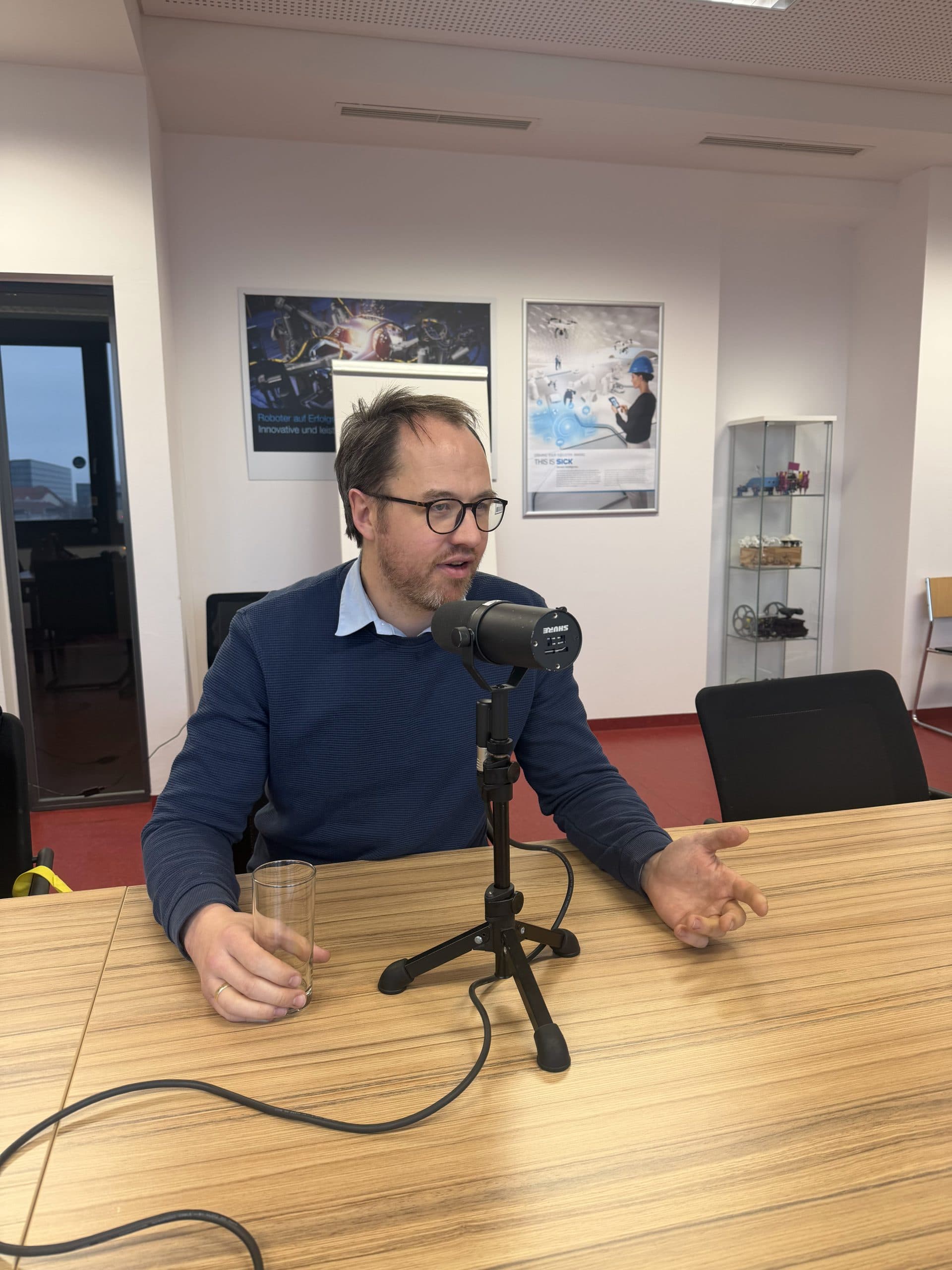 Ein Mann mit Brille und blauem Pullover sitzt an einem Tisch und spricht in ein Mikrofon auf einem Ständer. Er hält ein Glas Wasser in der Hand und gestikuliert mit der anderen Hand. Im Hintergrund sind Plakate und Regale zu sehen.