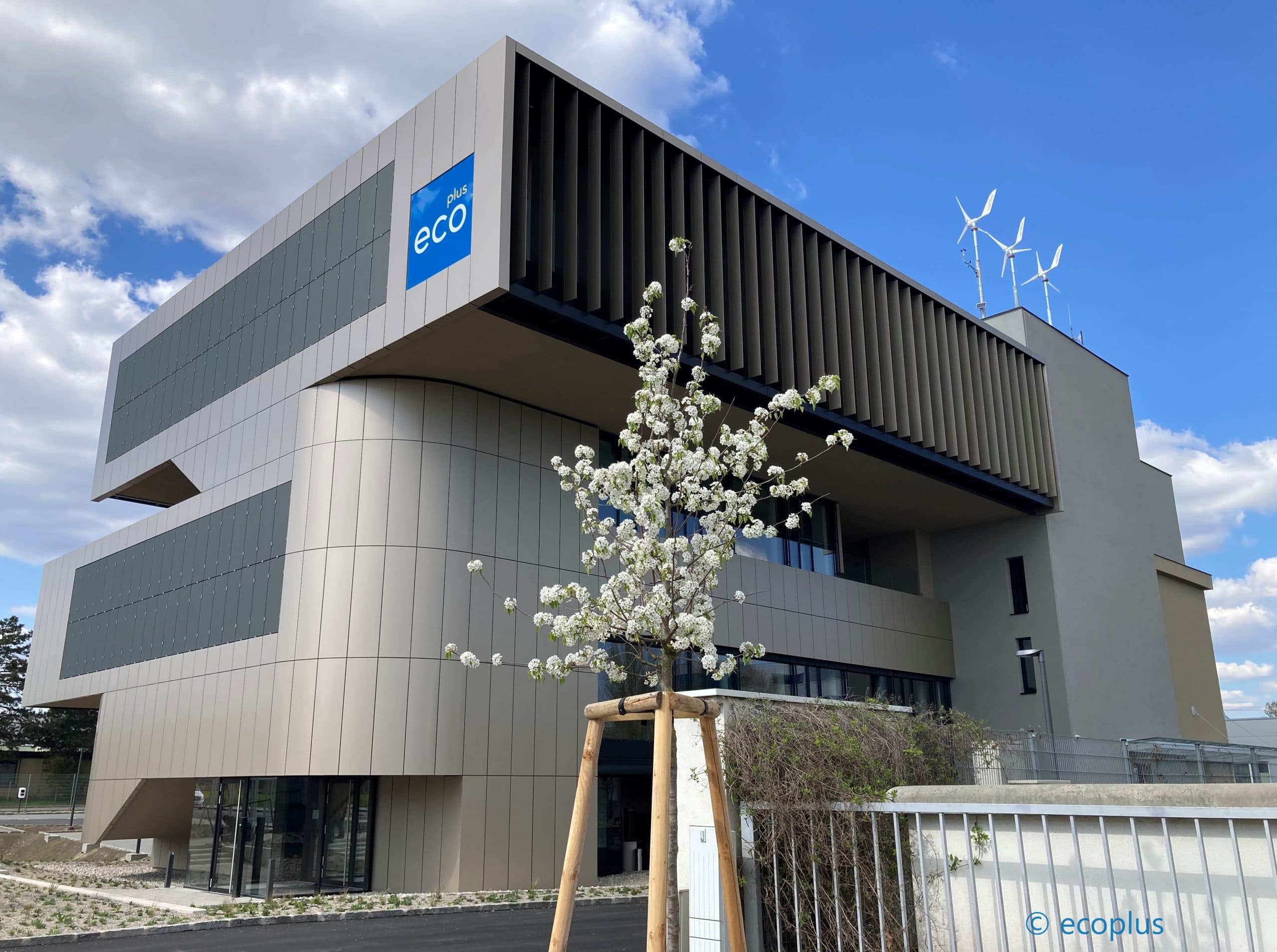 Ein modernes Gebäude mit einem blauen Ökozeichen, beigen und dunkelgrauen Paneelen und Windturbinen auf dem Dach steht hinter einem blühenden Baum mit weißen Blumen unter einem teilweise bewölkten Himmel.