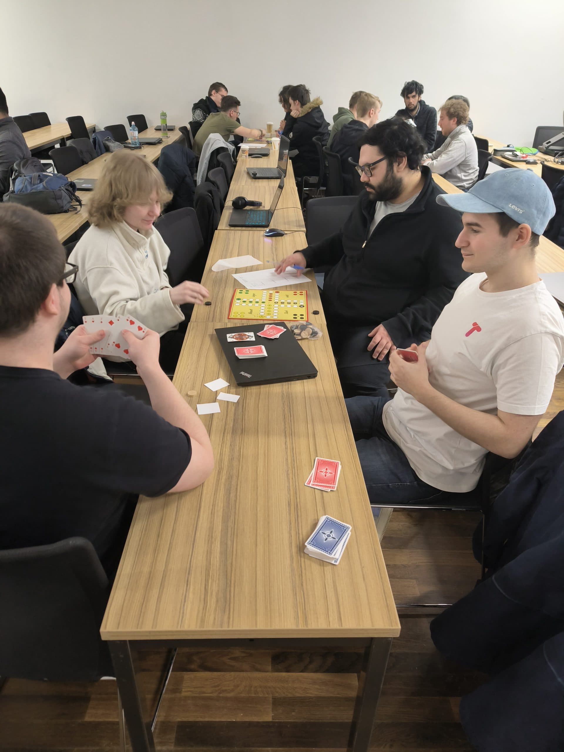 Vier Personen sitzen um einen Tisch in einem Klassenzimmer und spielen ein Karten- und Brettspiel, während andere im Hintergrund ebenfalls an Tischen sitzen. Der Raum hat einen Holzboden und eine weiße Wand.
